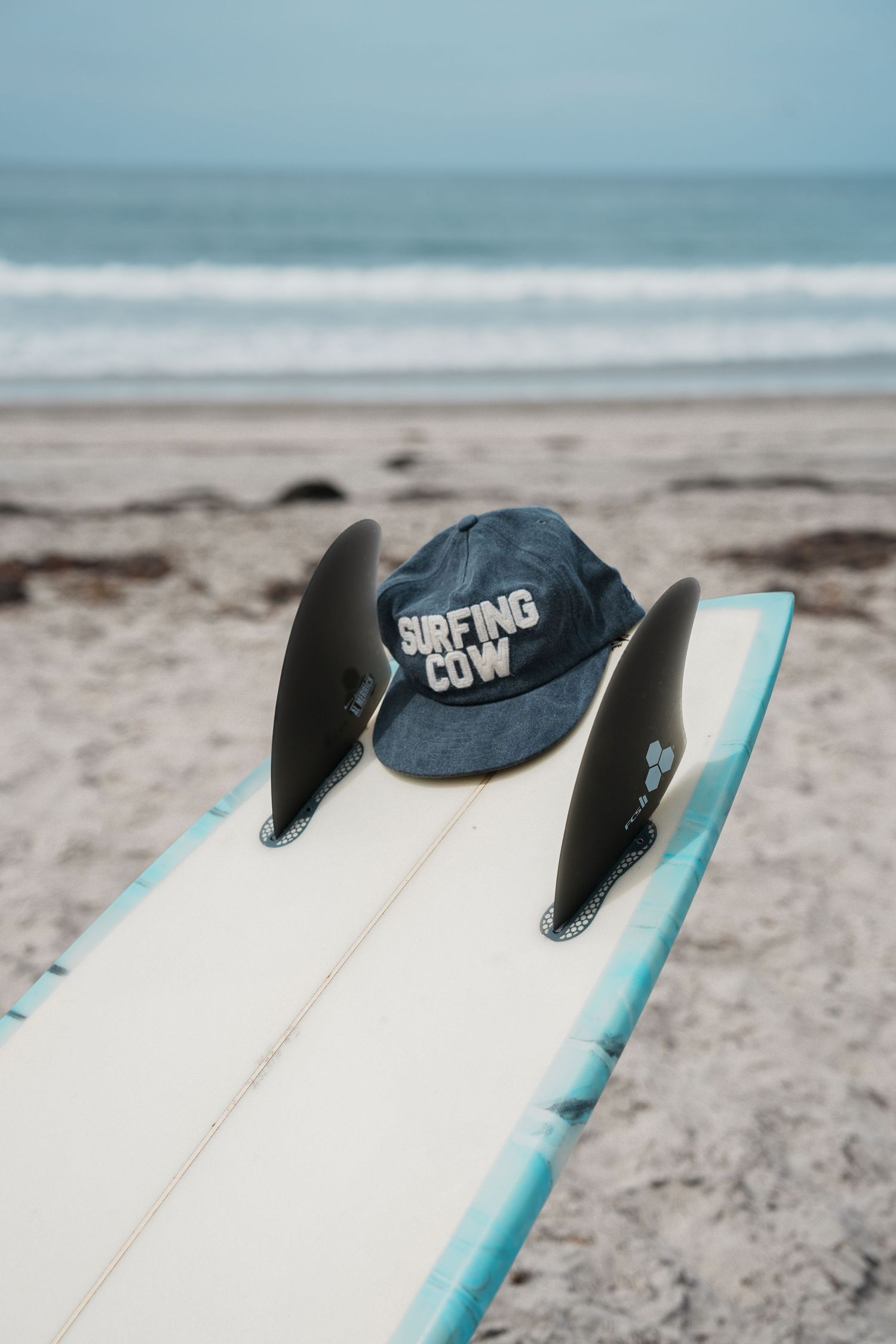 White surfboard with black fins rests on sandy beach with ocean waves in background.