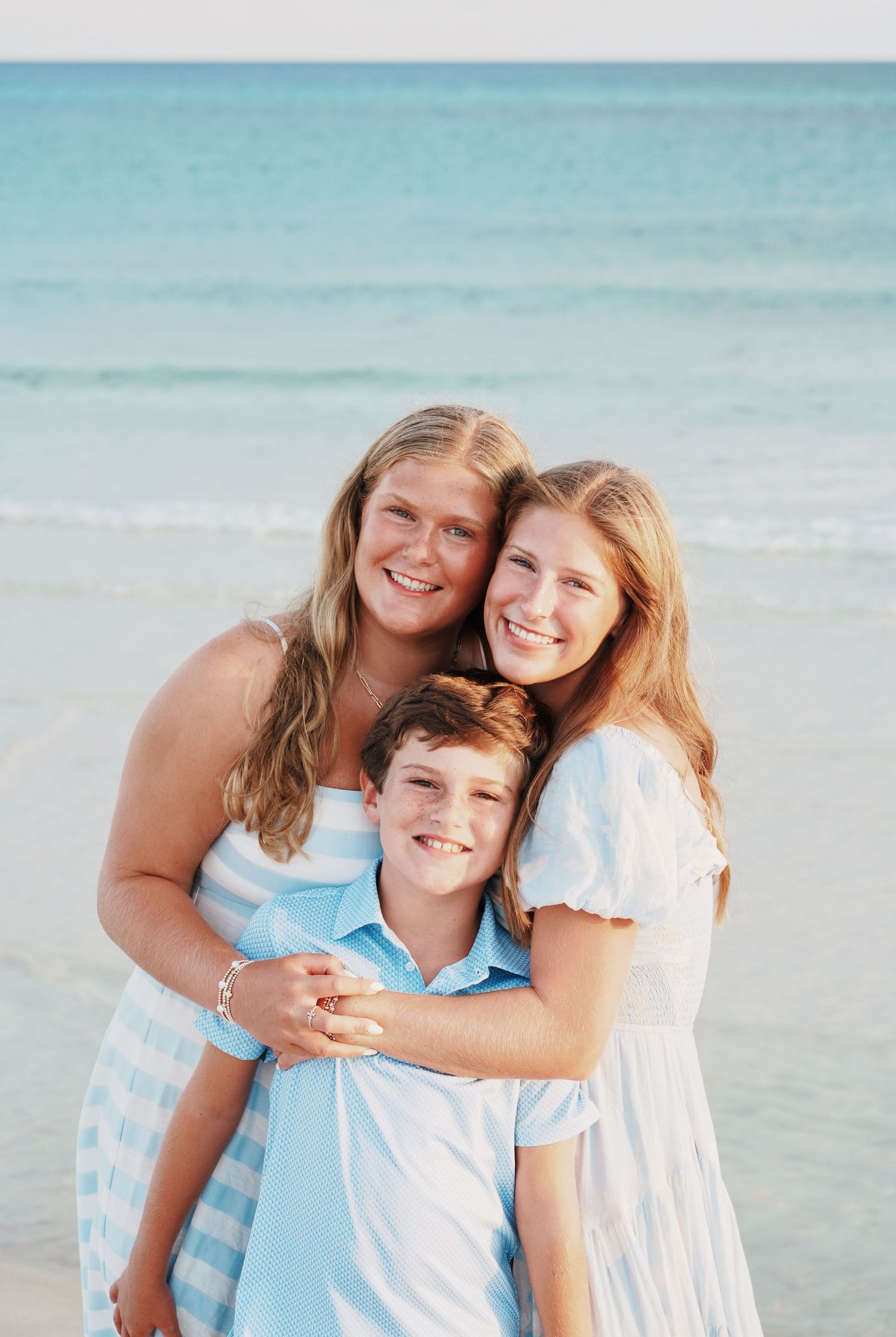 A family group dressed in light blue and white poses together for a beach portrait with turquoise ocean waters in background.