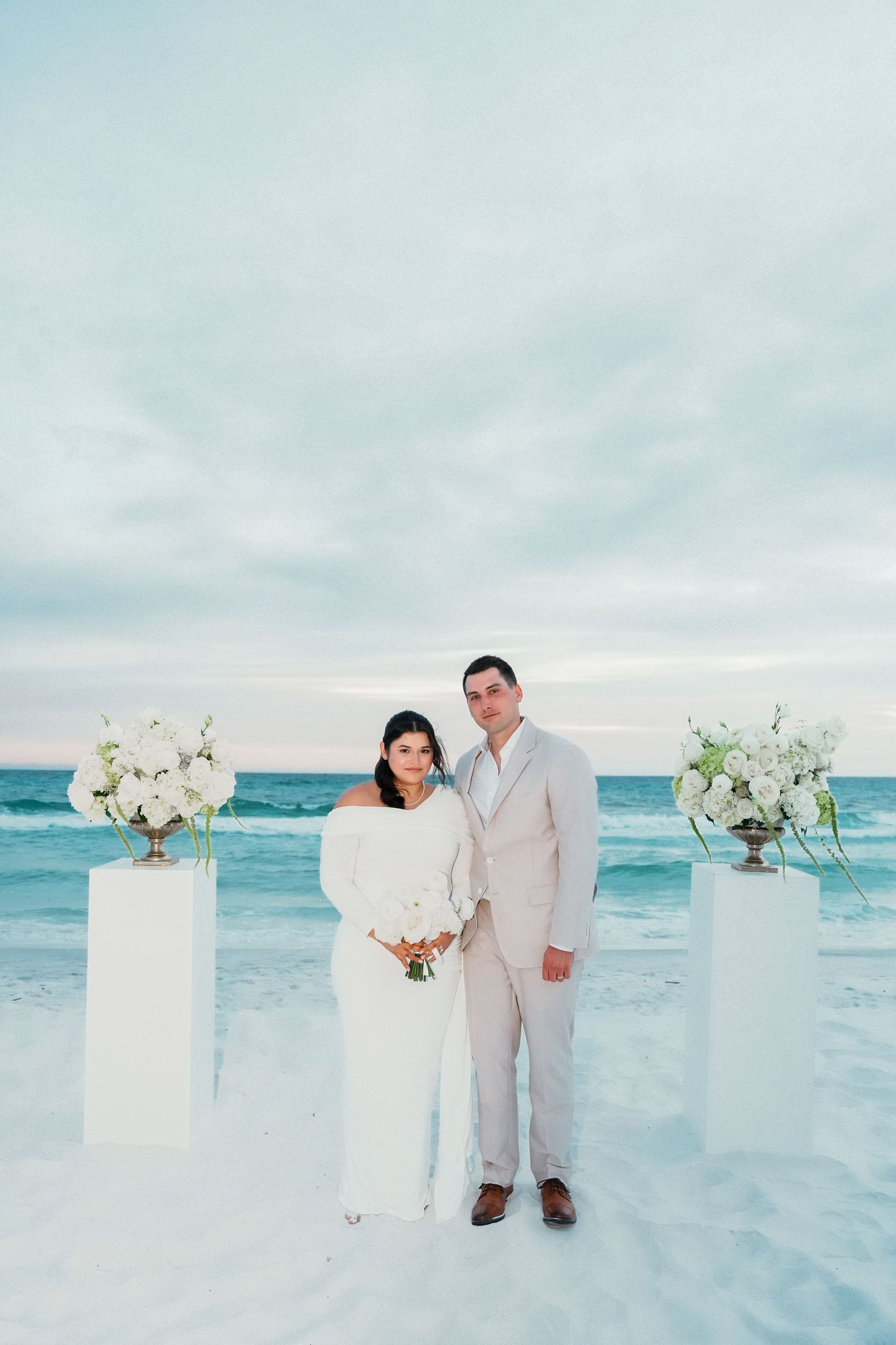 A couple stands on a white sandy beach with turquoise waters and floral arrangements on pedestals for their destination wedding.