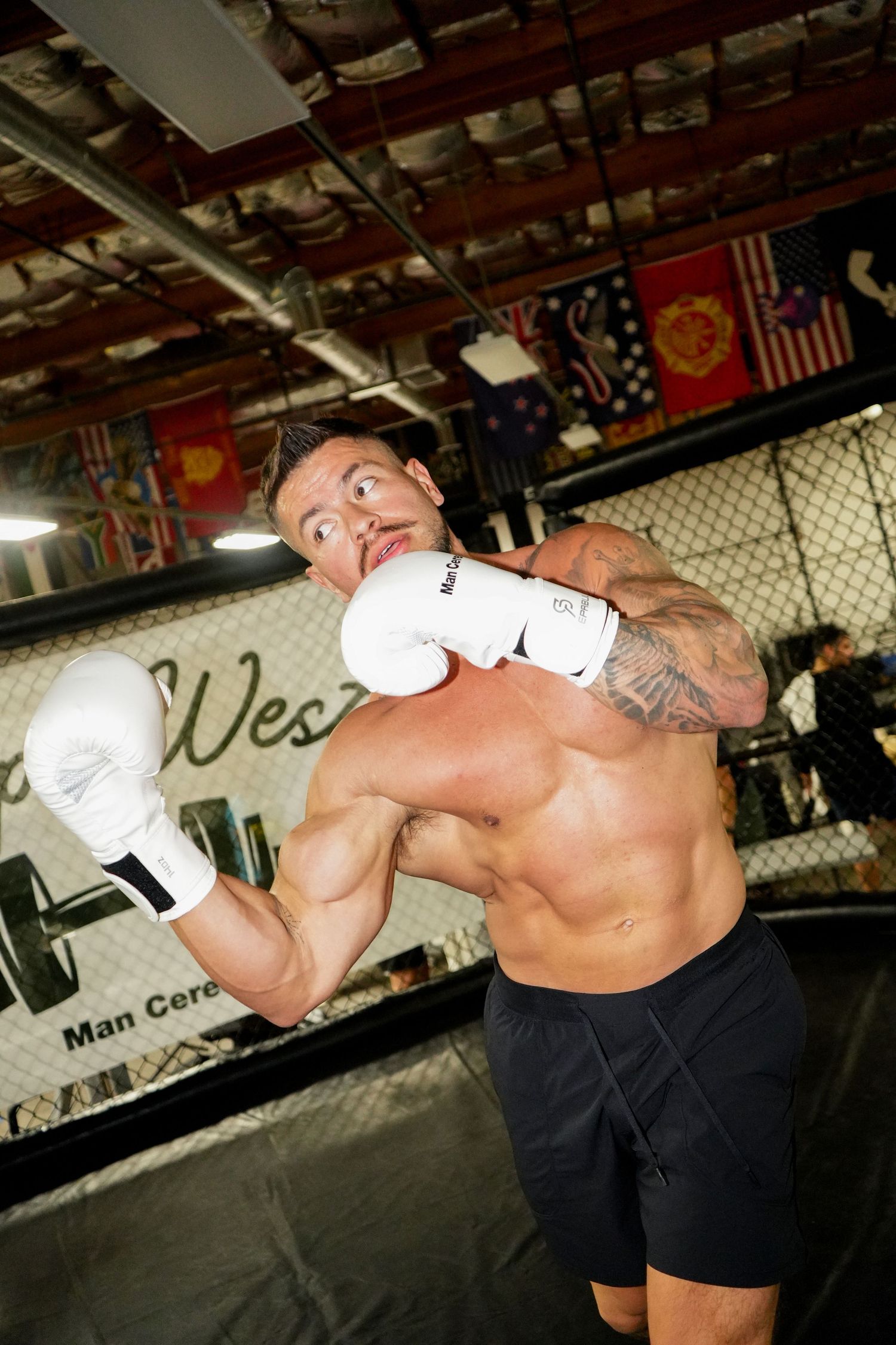 Athletic boxer training with white gloves in a gym with international flags hanging from the ceiling.