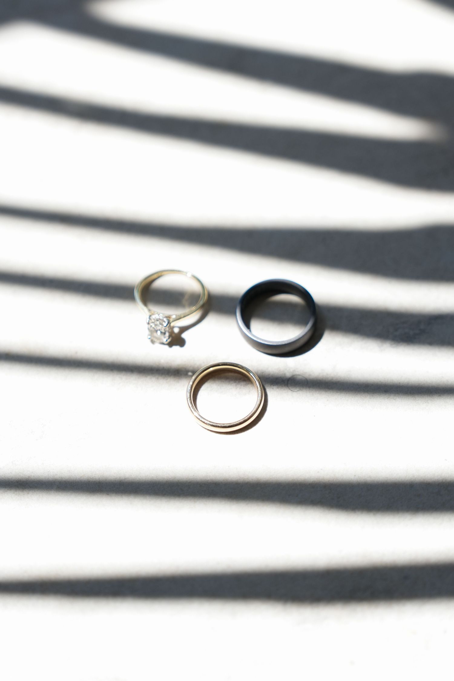 Three wedding rings arranged on a white surface with dramatic zebra-stripe shadows, including a diamond engagement ring.