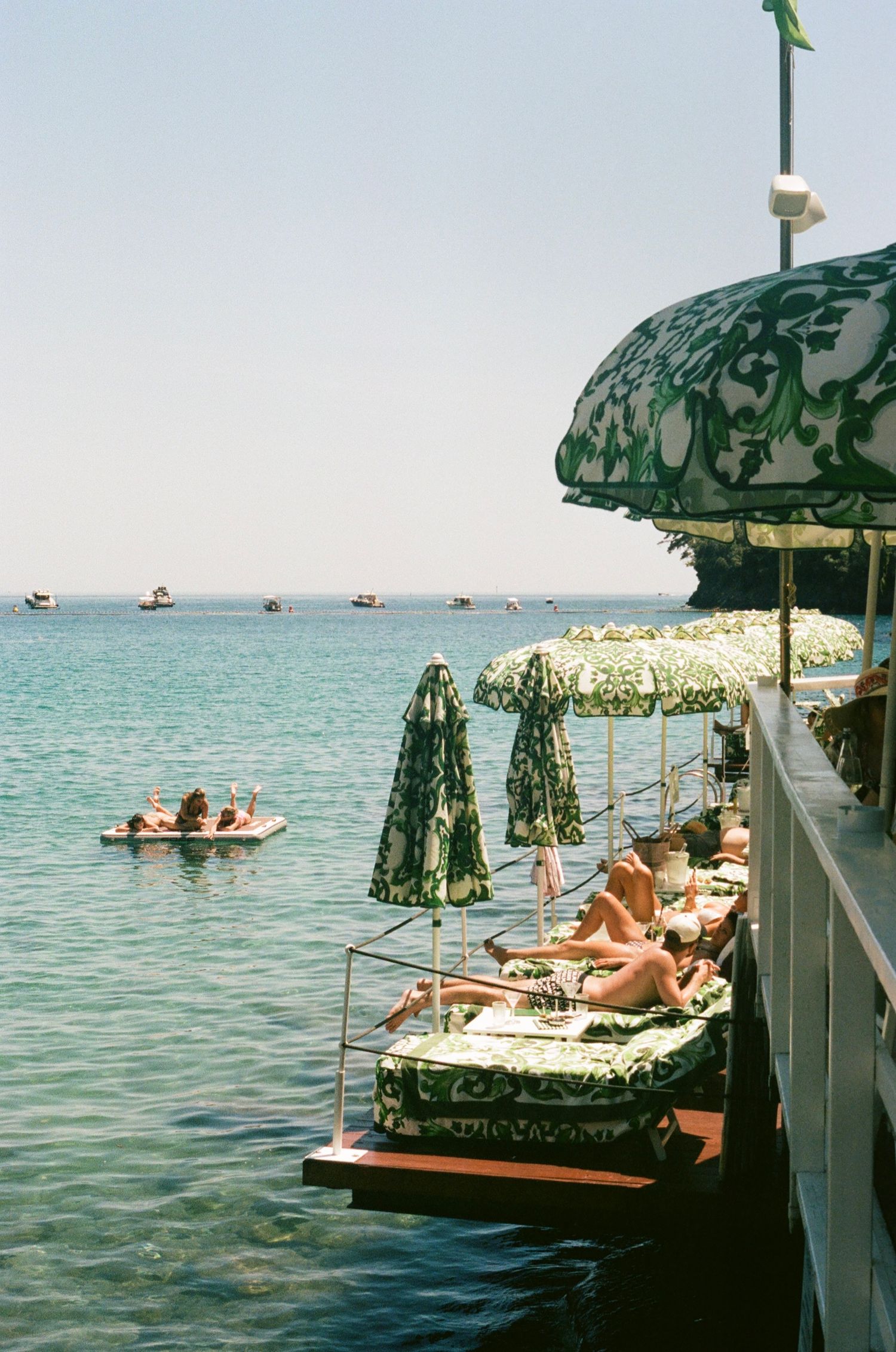 Sunbathers relax on elegant green-patterned daybeds by the sea, with matching umbrellas and boats in the calm blue water.