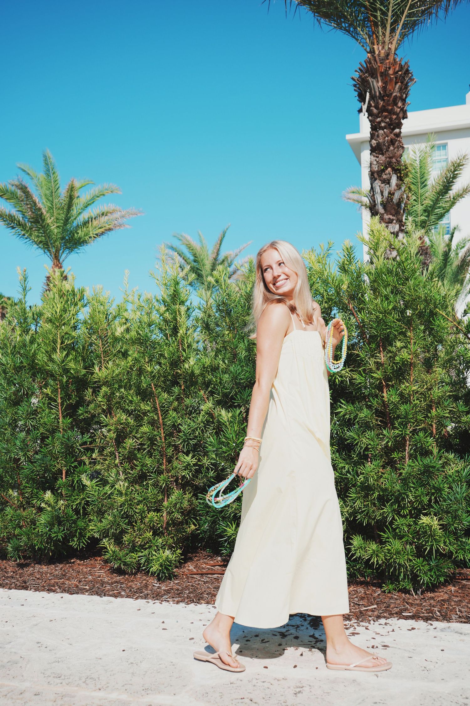 A person in a white summer dress walks past palm trees and greenery on a sunny day at a tropical resort.