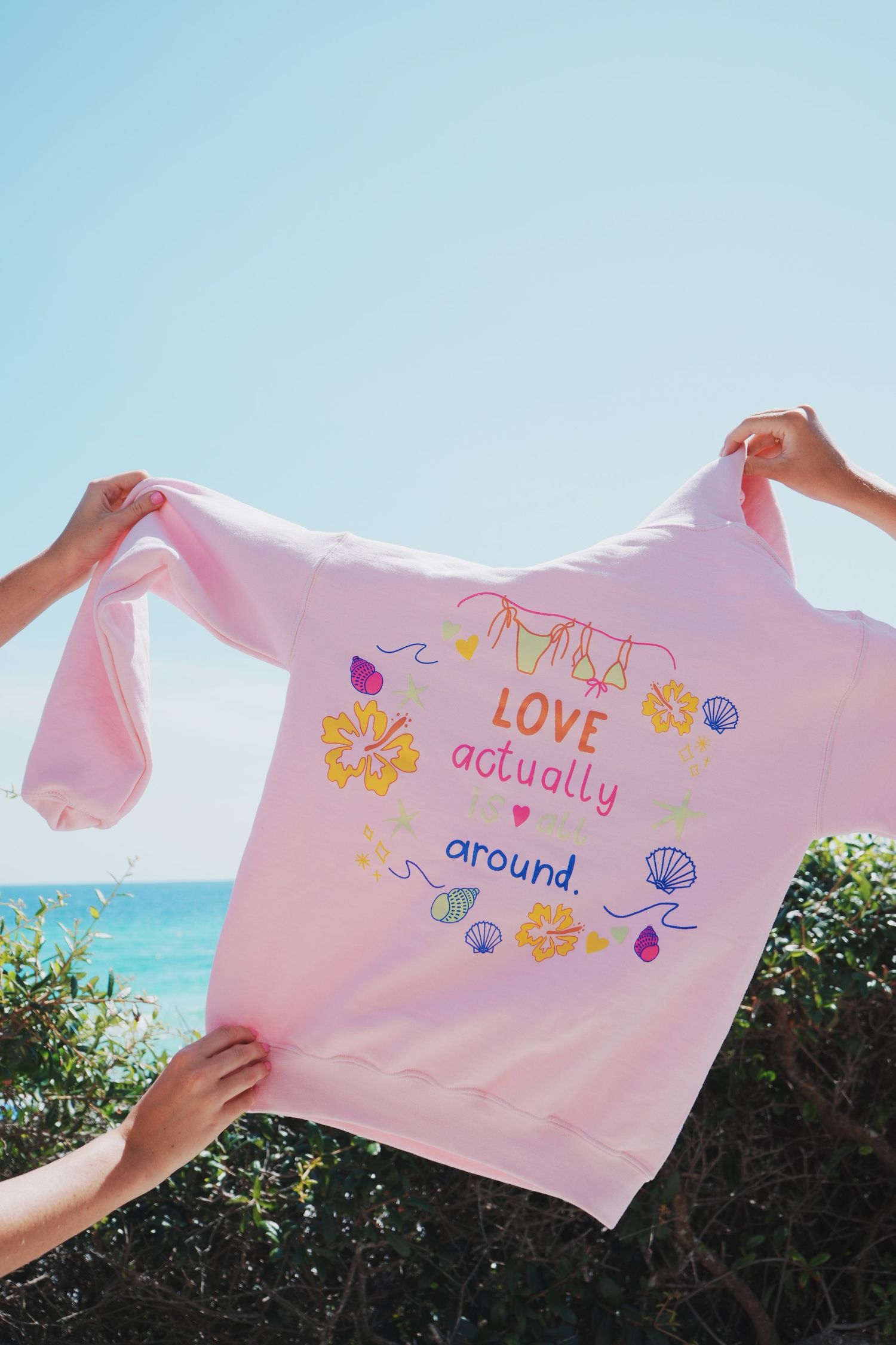 Hands holding up a pink sweatshirt with 'Love actually around' text and decorative designs against blue sky and ocean.