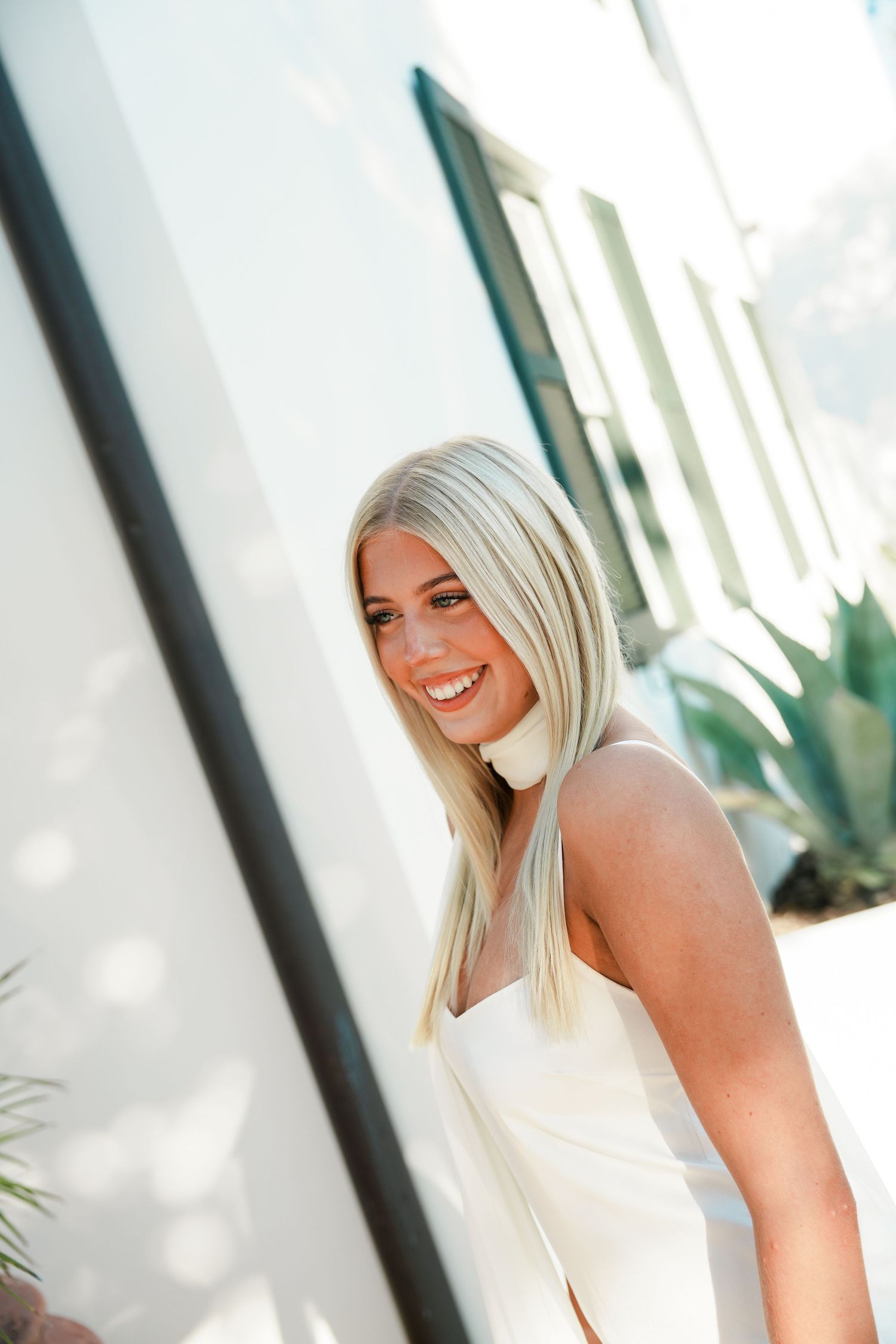 Portrait in soft natural lighting shows someone with platinum blonde hair wearing a white sleeveless top and a bright smile.