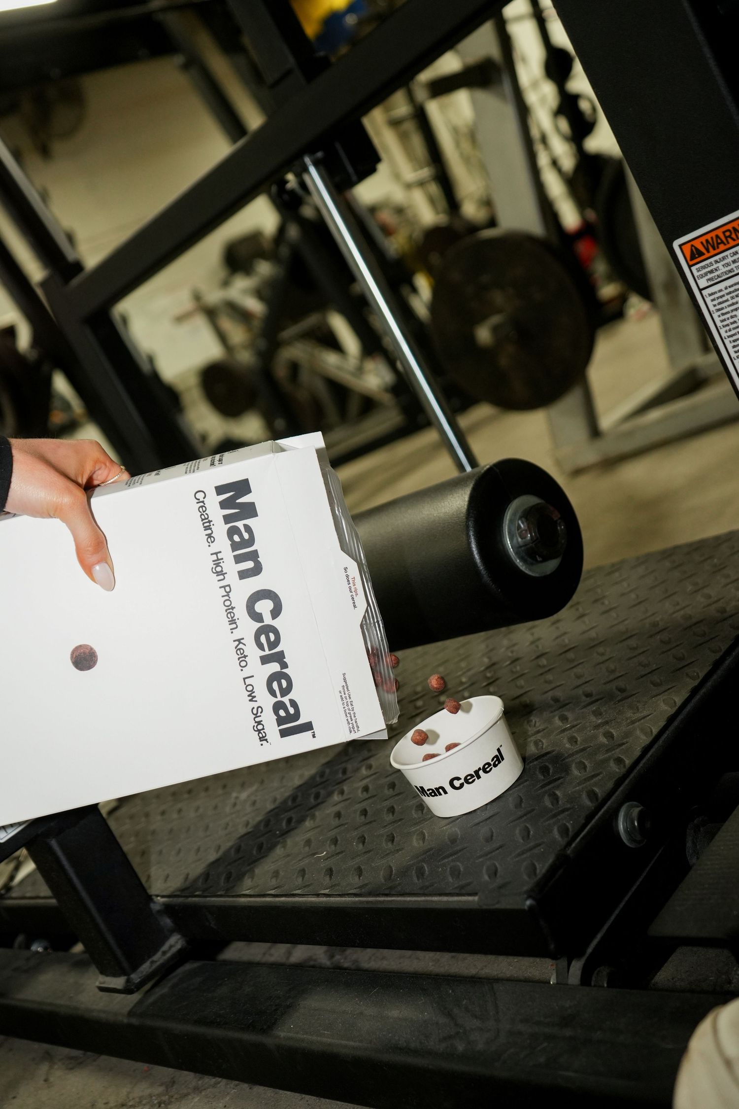Close-up shot of someone holding a Man Cereal product package next to gym equipment.