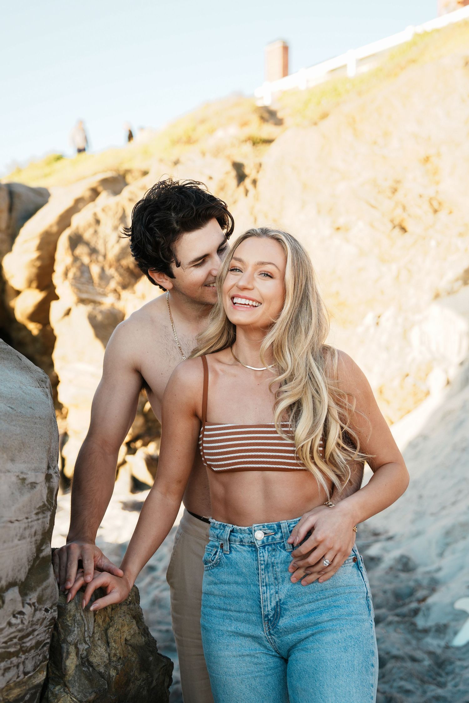 A romantic couple poses together on a beach with rocky cliffs during golden hour sunset.