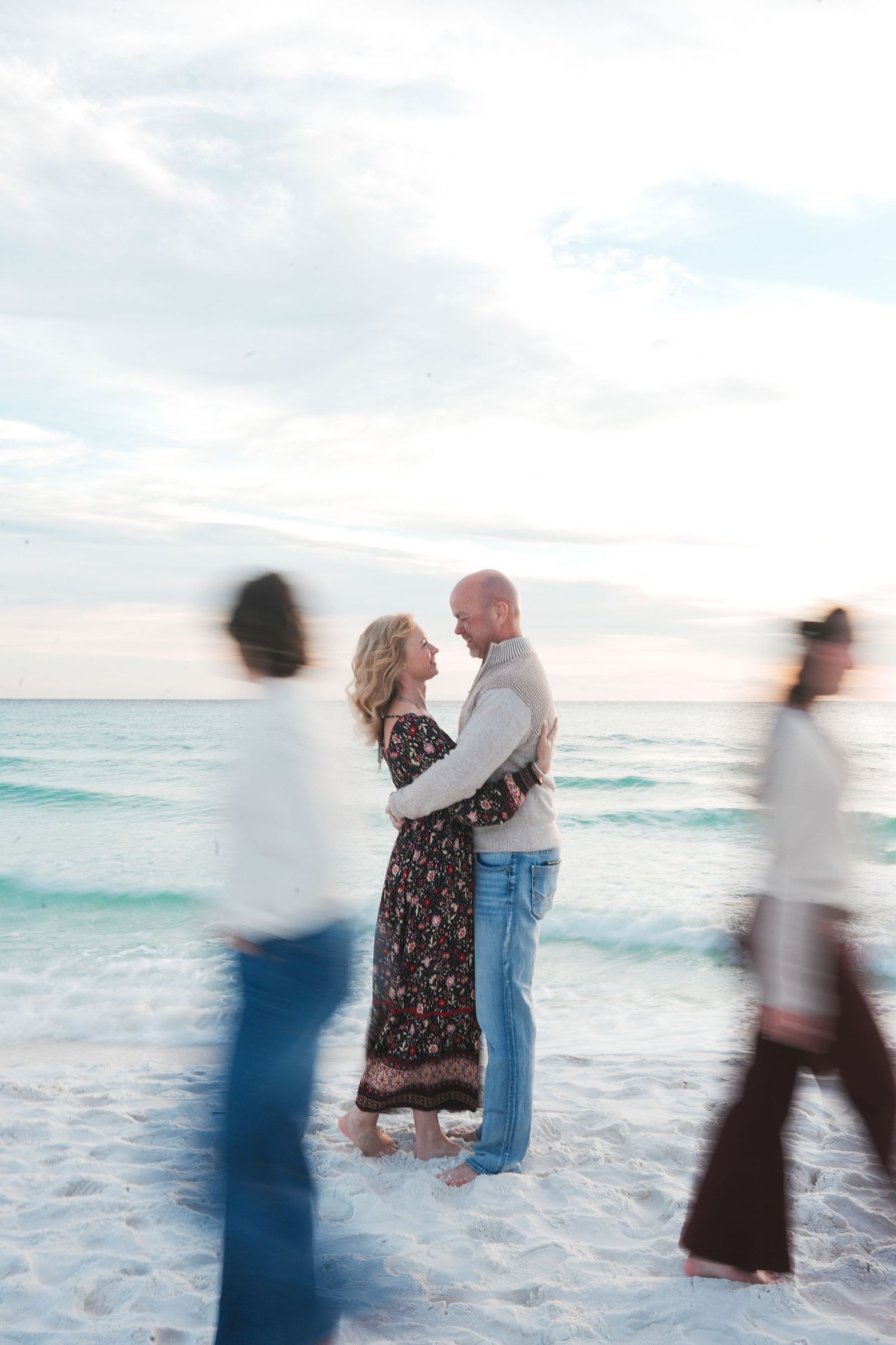 A couple shares a romantic moment together at sunset on a beach with blurred motion figures walking past in the background.