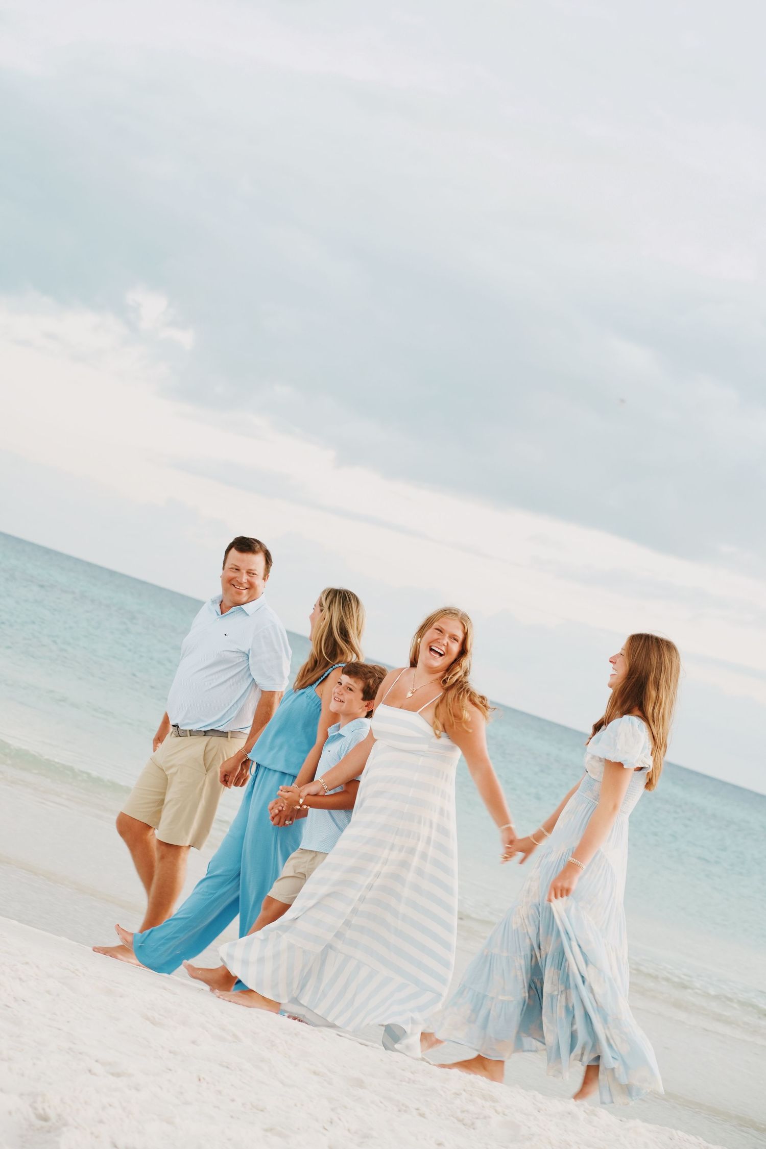 A family in coordinating blue and white outfits enjoys a playful moment together while walking on a sunny beach.