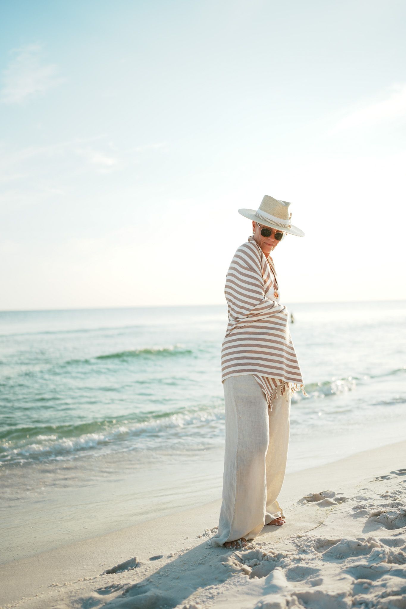 A person in a striped outfit and sun hat walks along a serene beach at sunset with waves rolling in.