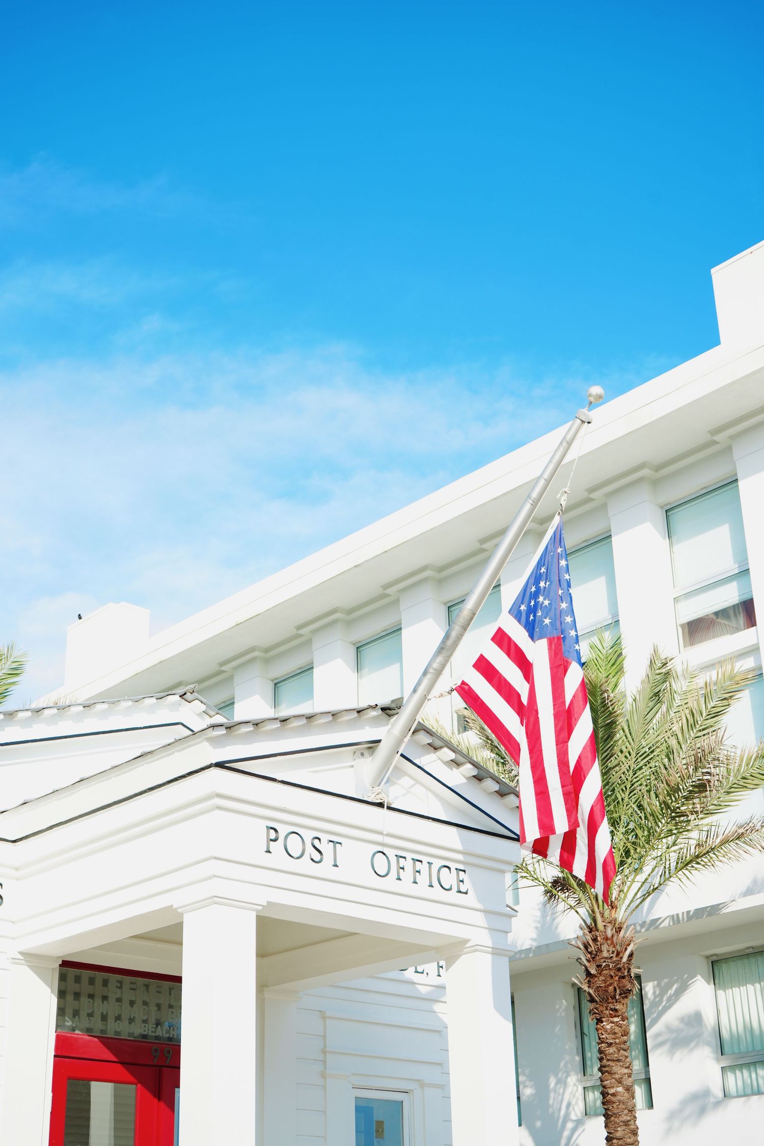 White post office building with American flags and palm trees under bright blue sky in beach town setting.