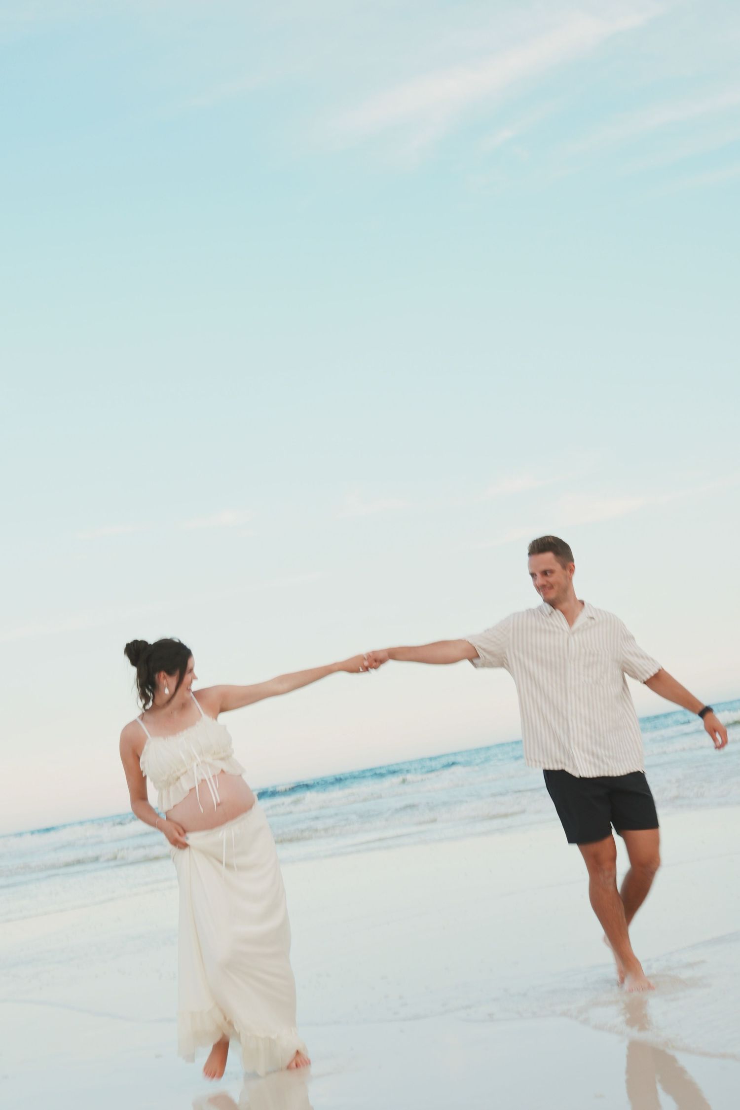 Couple in white outfits playfully dancing together on a sunny beach with ocean waves in the background.