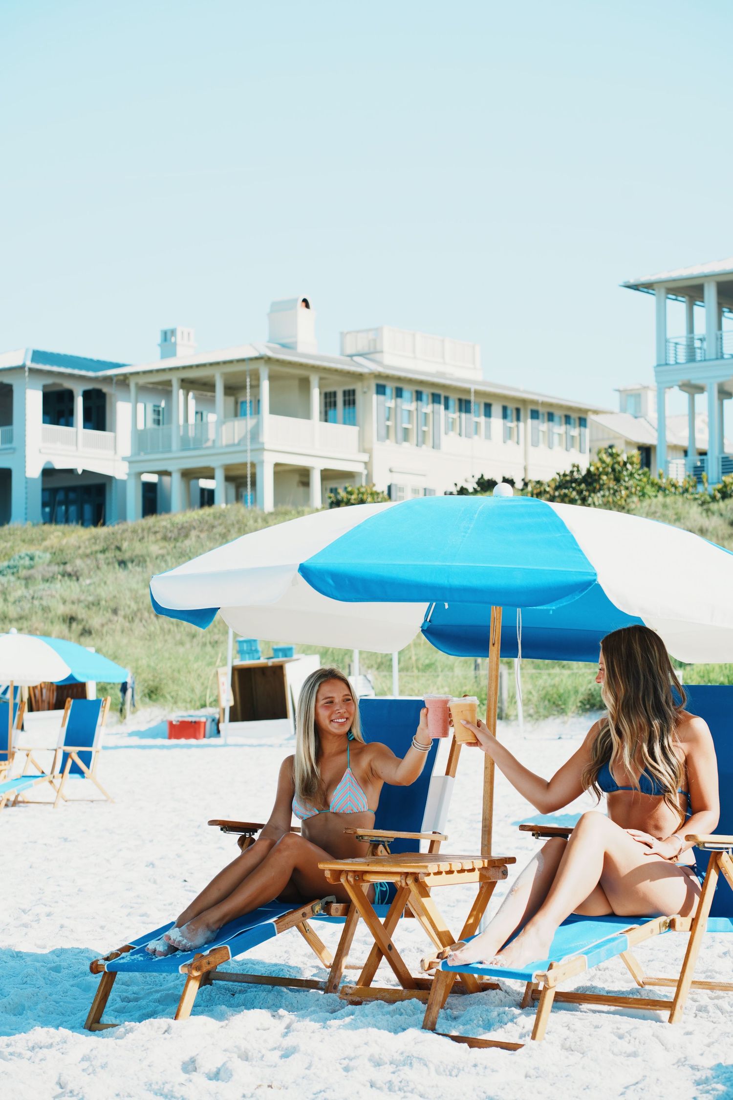 Group lounging on beach chairs with umbrellas while enjoying drinks by coastal homes.