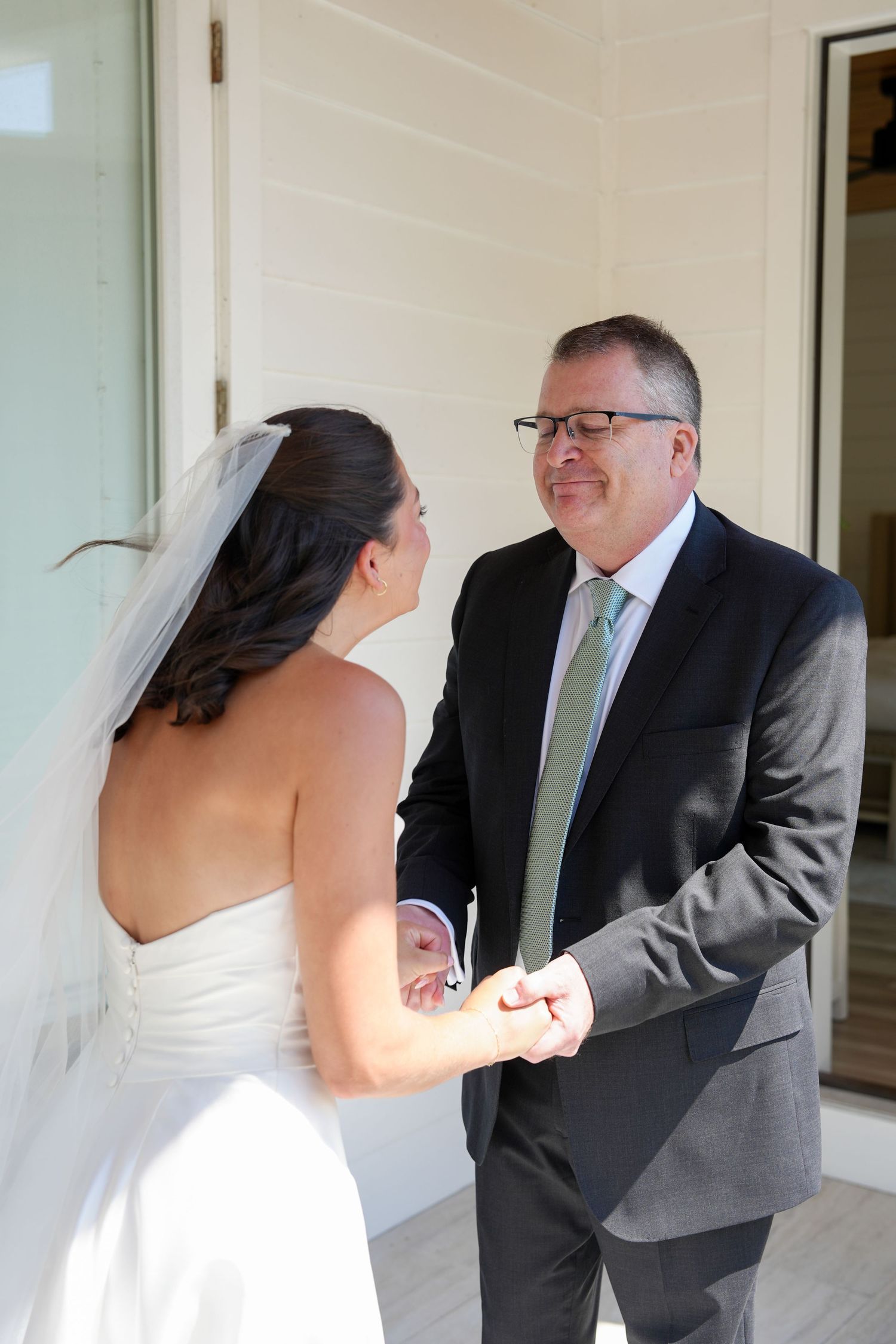 A bride in a white gown holds hands with a man in a gray suit and green tie during a wedding first look.