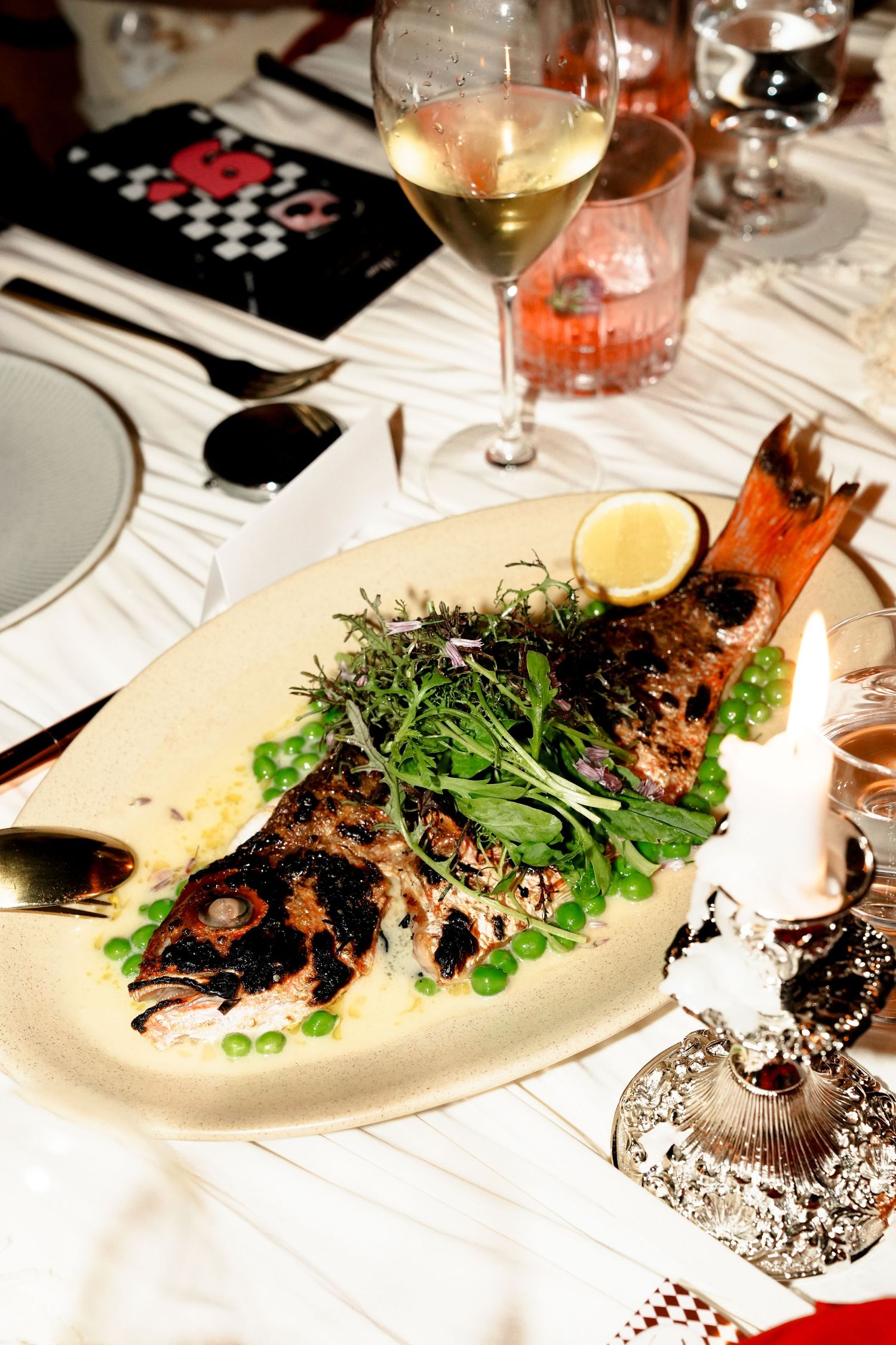 Whole roasted fish served on platter with peas, herbs, lemon, and white wine at elegant dinner.