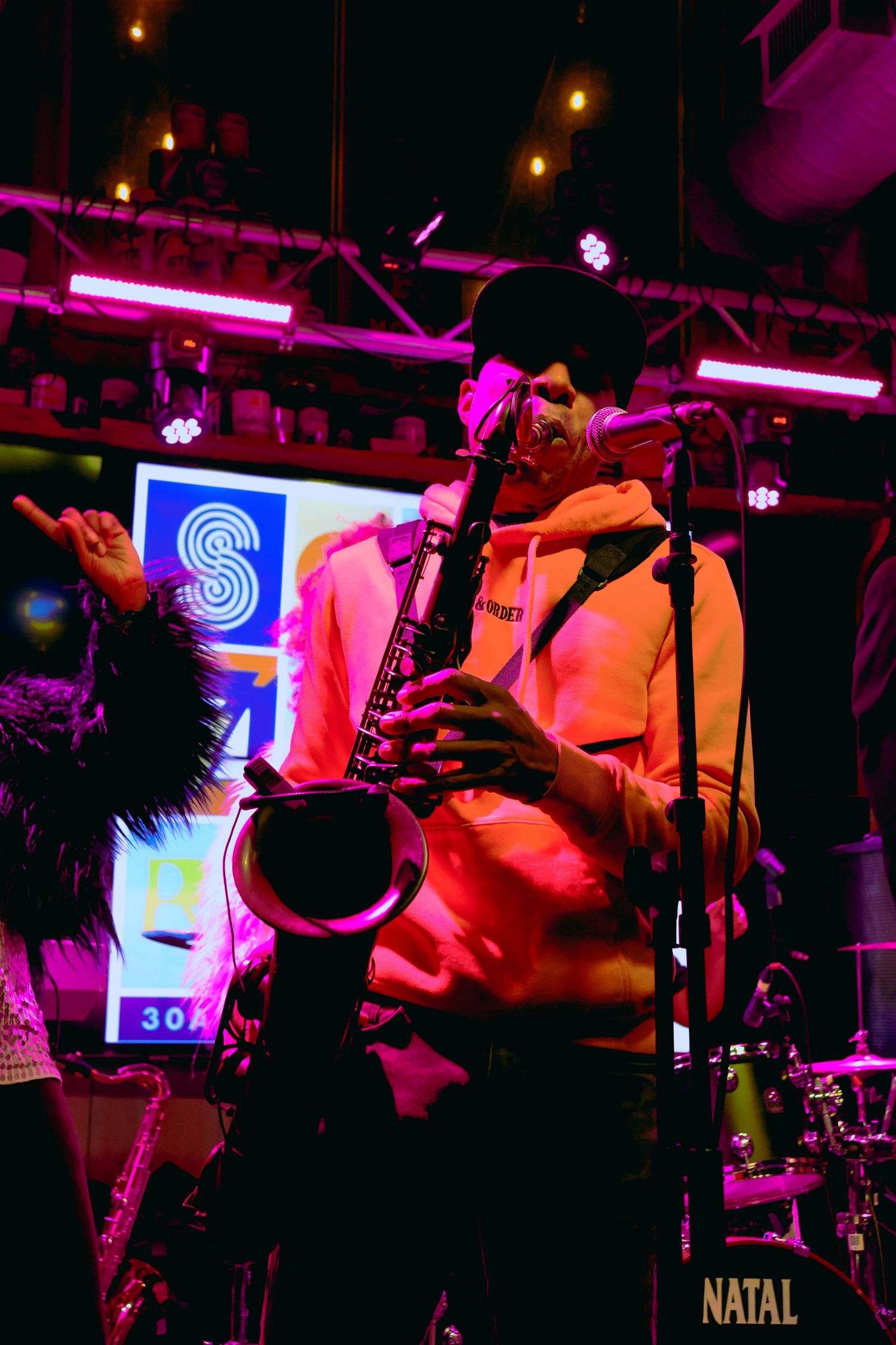 Saxophone player performing under vibrant pink and purple stage lights near neon signage.