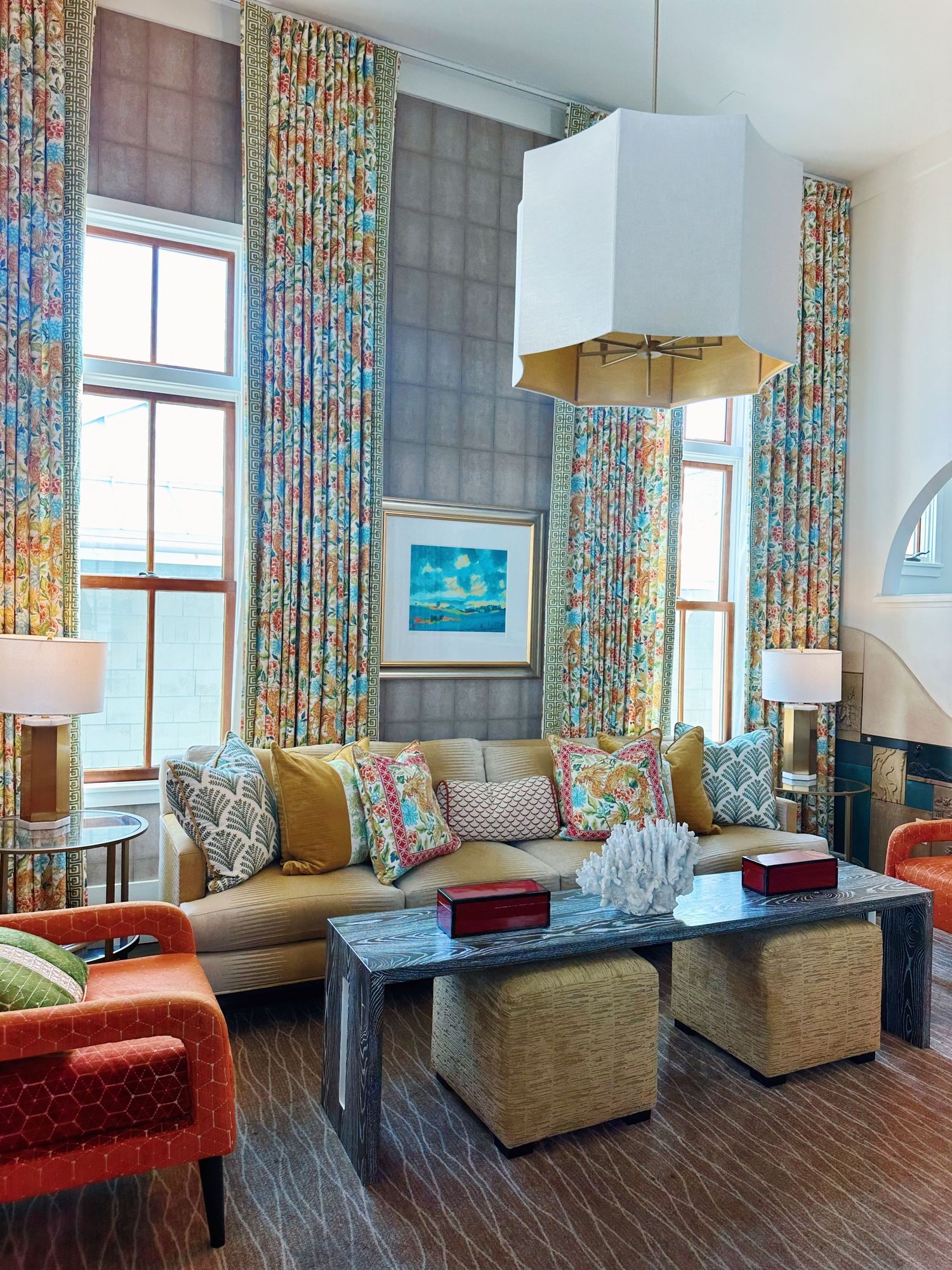 Modern coastal living room with floral curtains, beige sofa, orange accent chairs and hardwood floors showcases oceanfront views.
