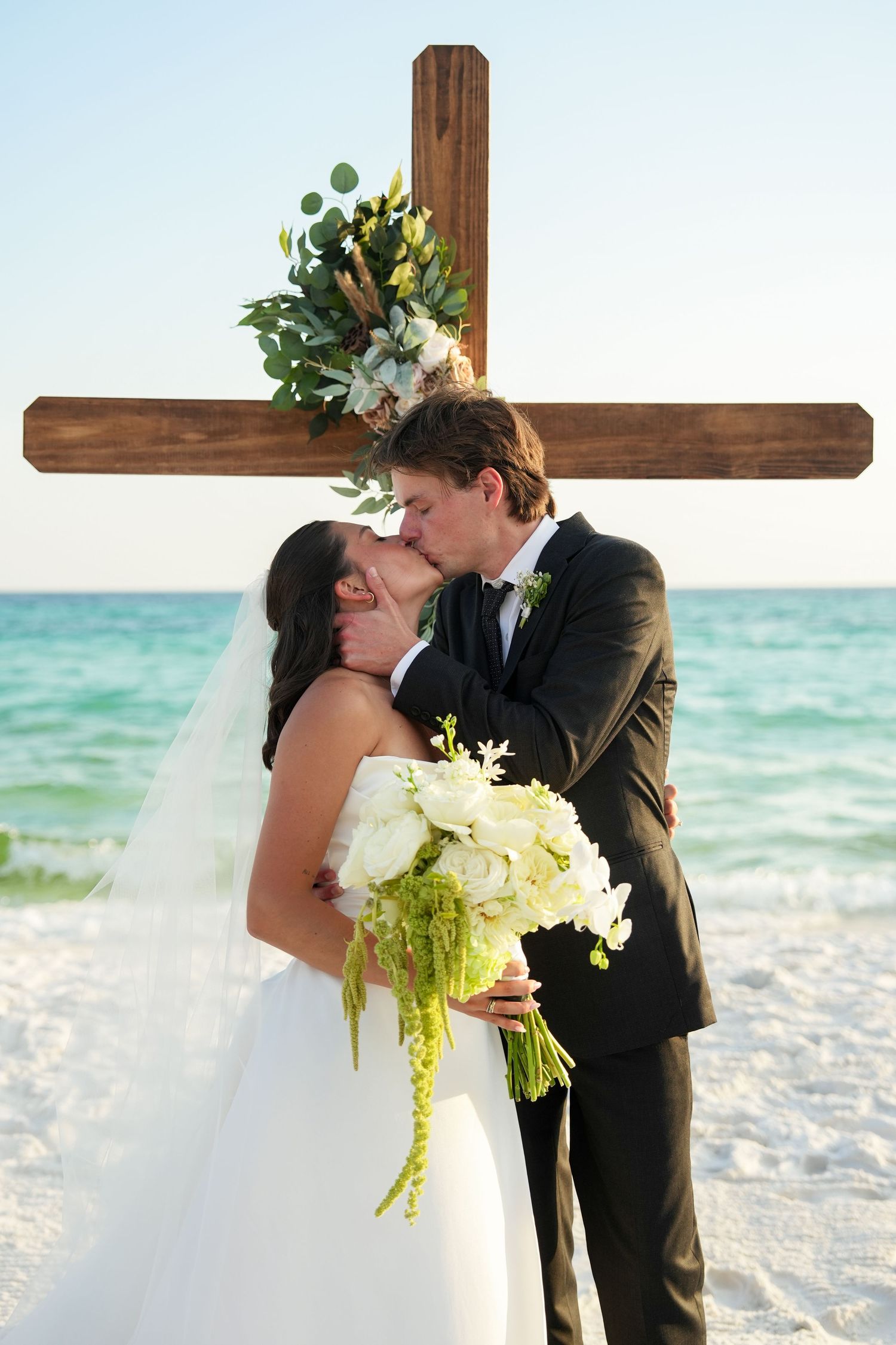 Bride and groom share first kiss at beach wedding ceremony before wooden cross decorated with white flowers.