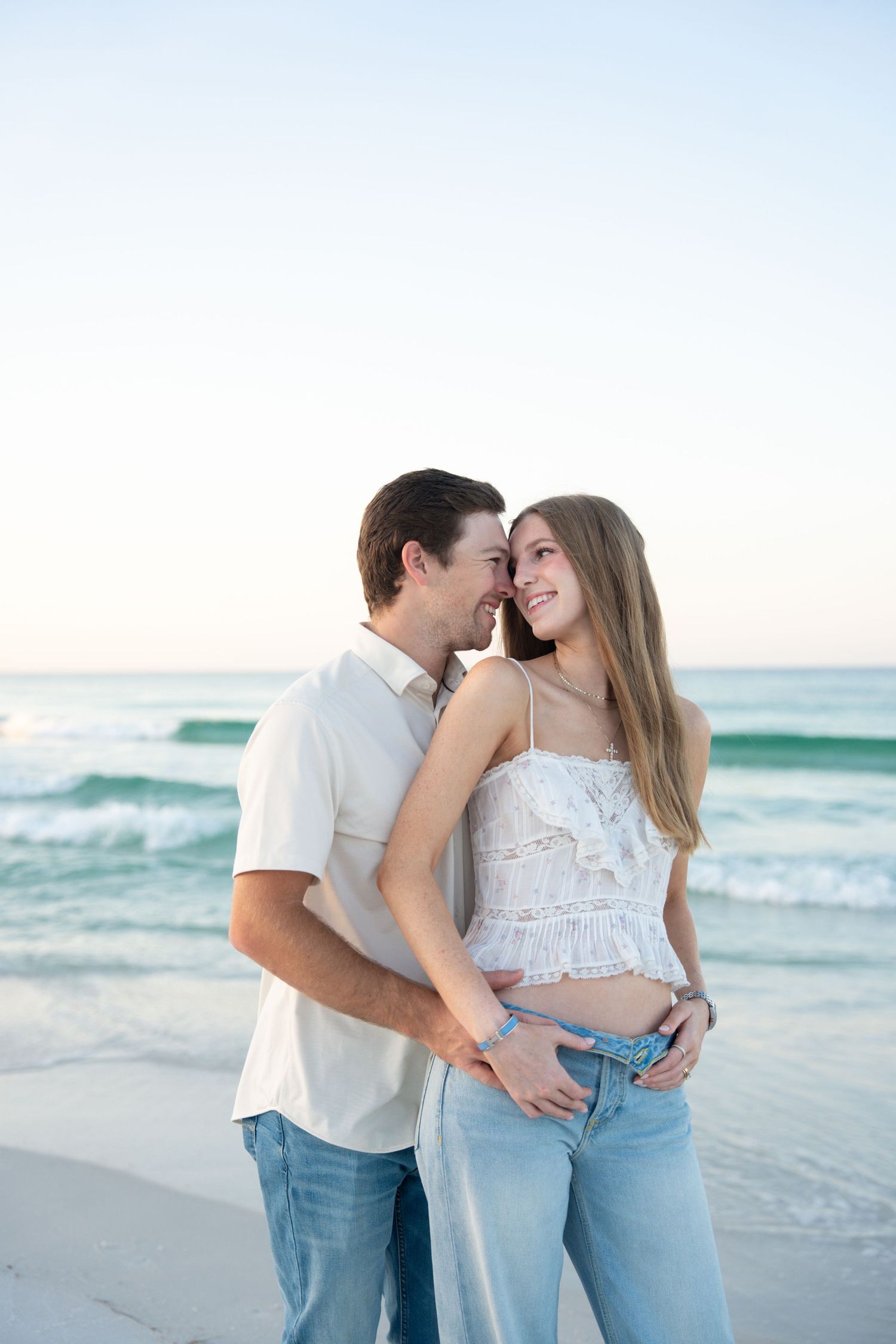 Romantic couple embracing on a beach at sunset, woman in white top and jeans smiling at partner near ocean waves.