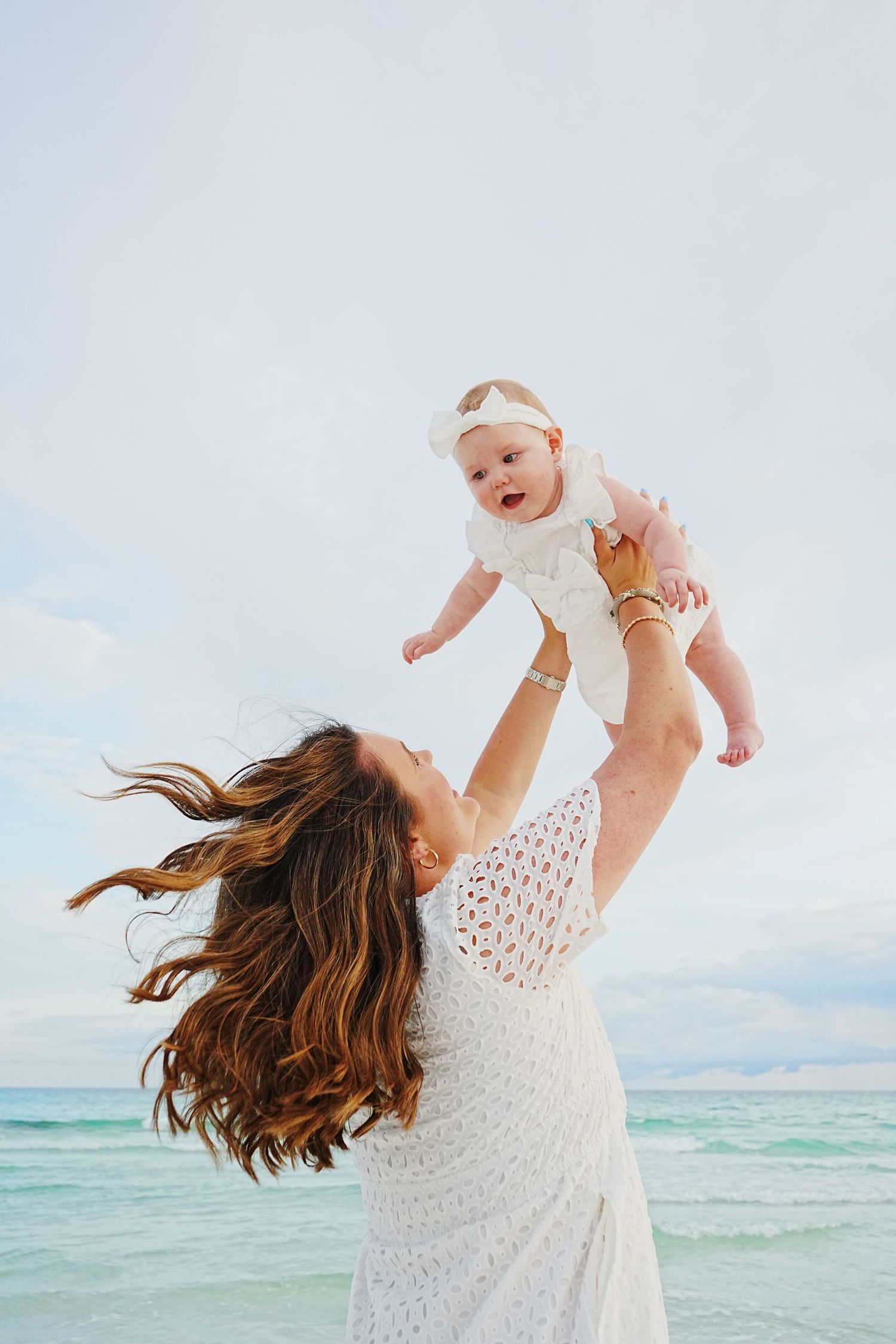 Joyful mom in white lace dress lifts laughing baby in air on beach with turquoise ocean and cloudy sky background.