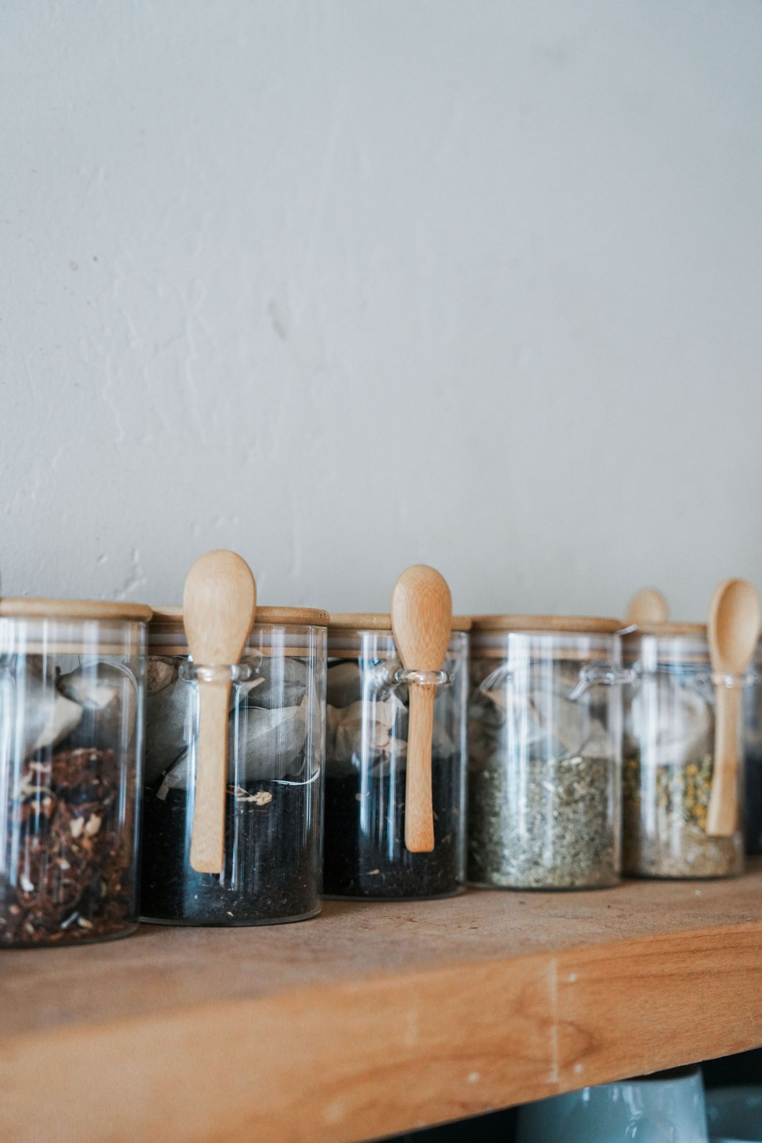 Glass jars filled with various spices and herbs arranged on a wooden shelf with wooden spoons.