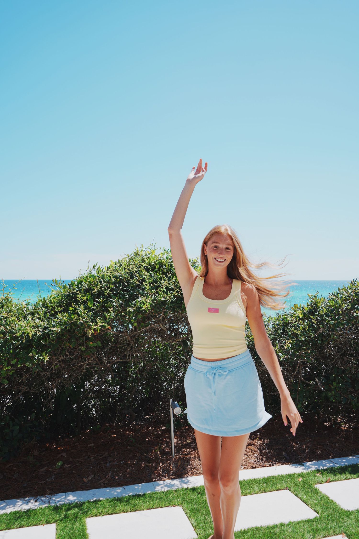 A person in summer clothing poses with one arm raised against a coastal backdrop.