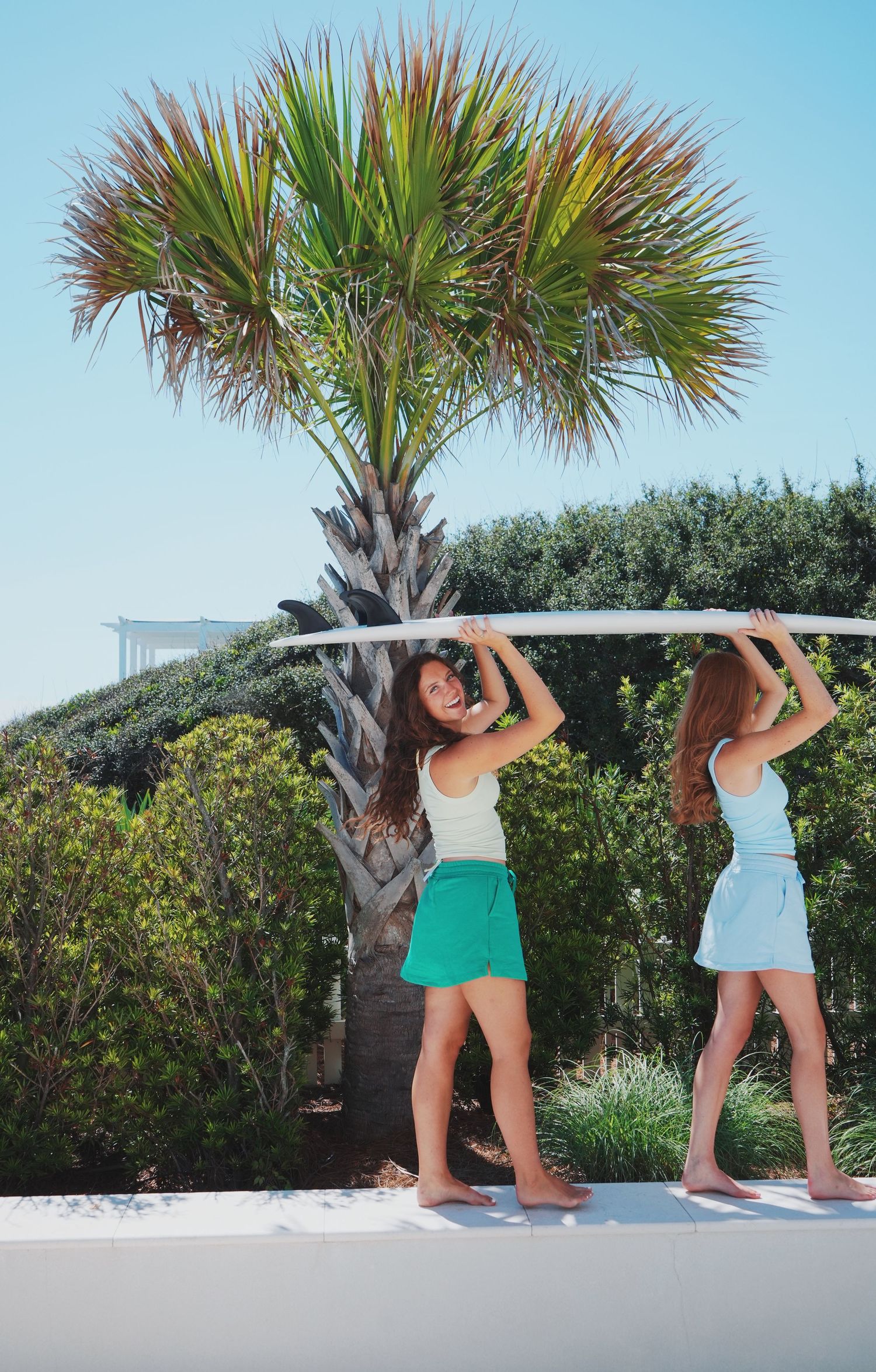 Two people carrying surfboard overhead beneath palm tree in coastal setting.
