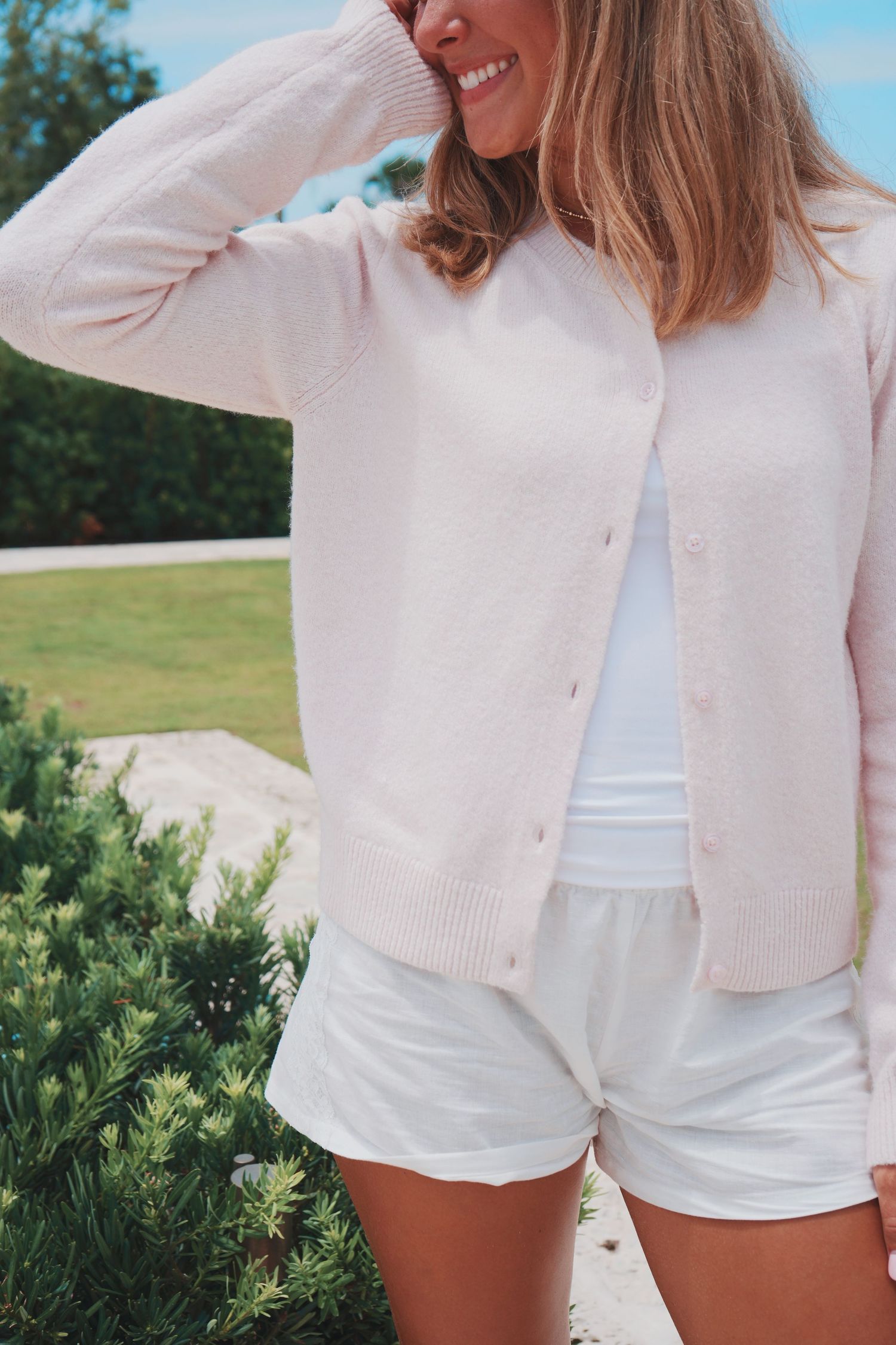 Summer fashion outfit with light pink cardigan, white tank top and white shorts against green garden backdrop.