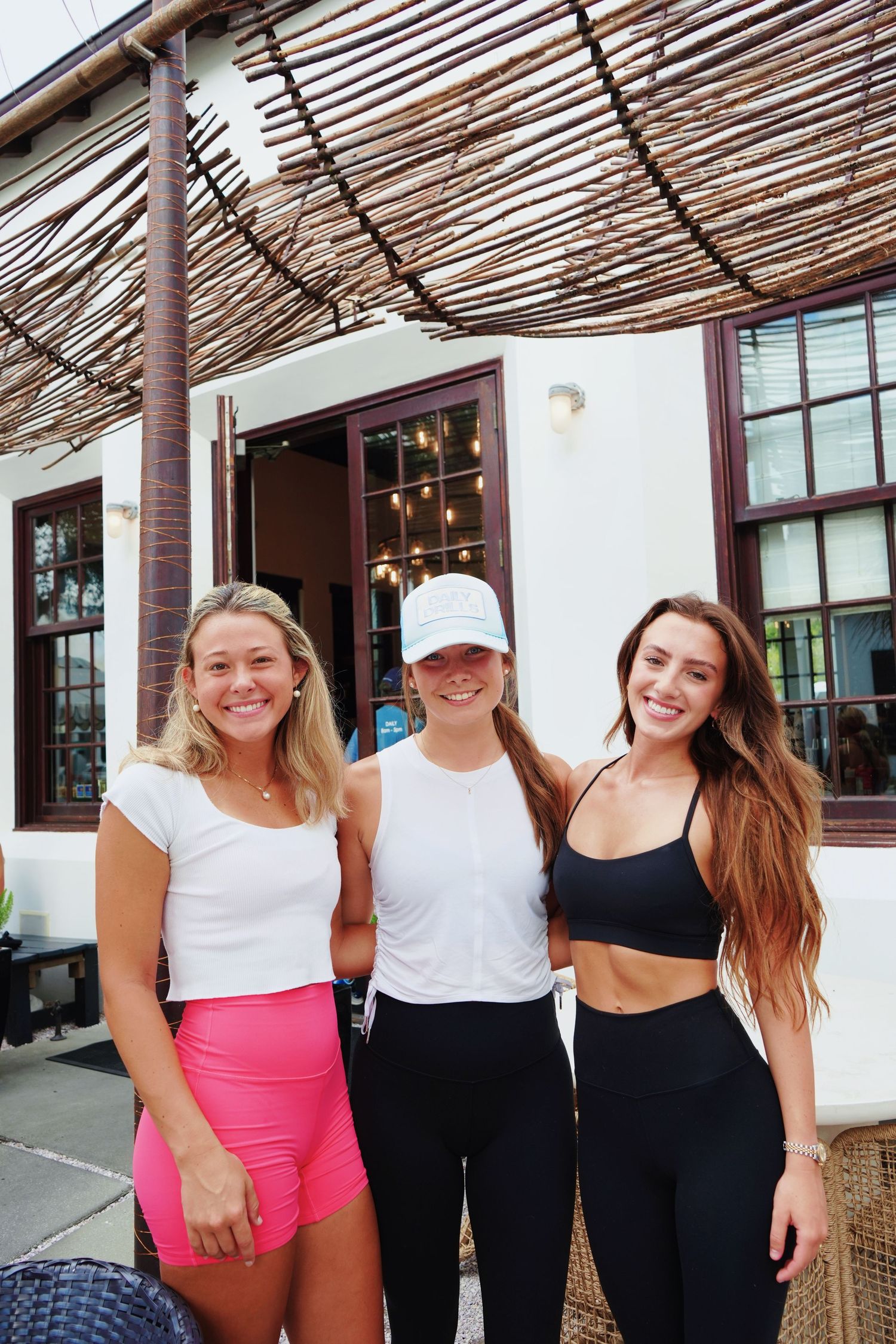 Three casual friends wearing summer outfits pose together under a covered outdoor patio area.