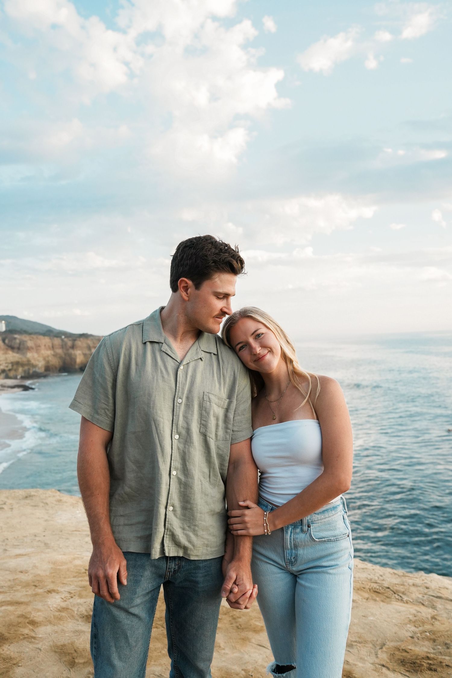 A couple stands together on a coastal cliff overlooking the ocean on a sunny day with blue skies and white clouds.