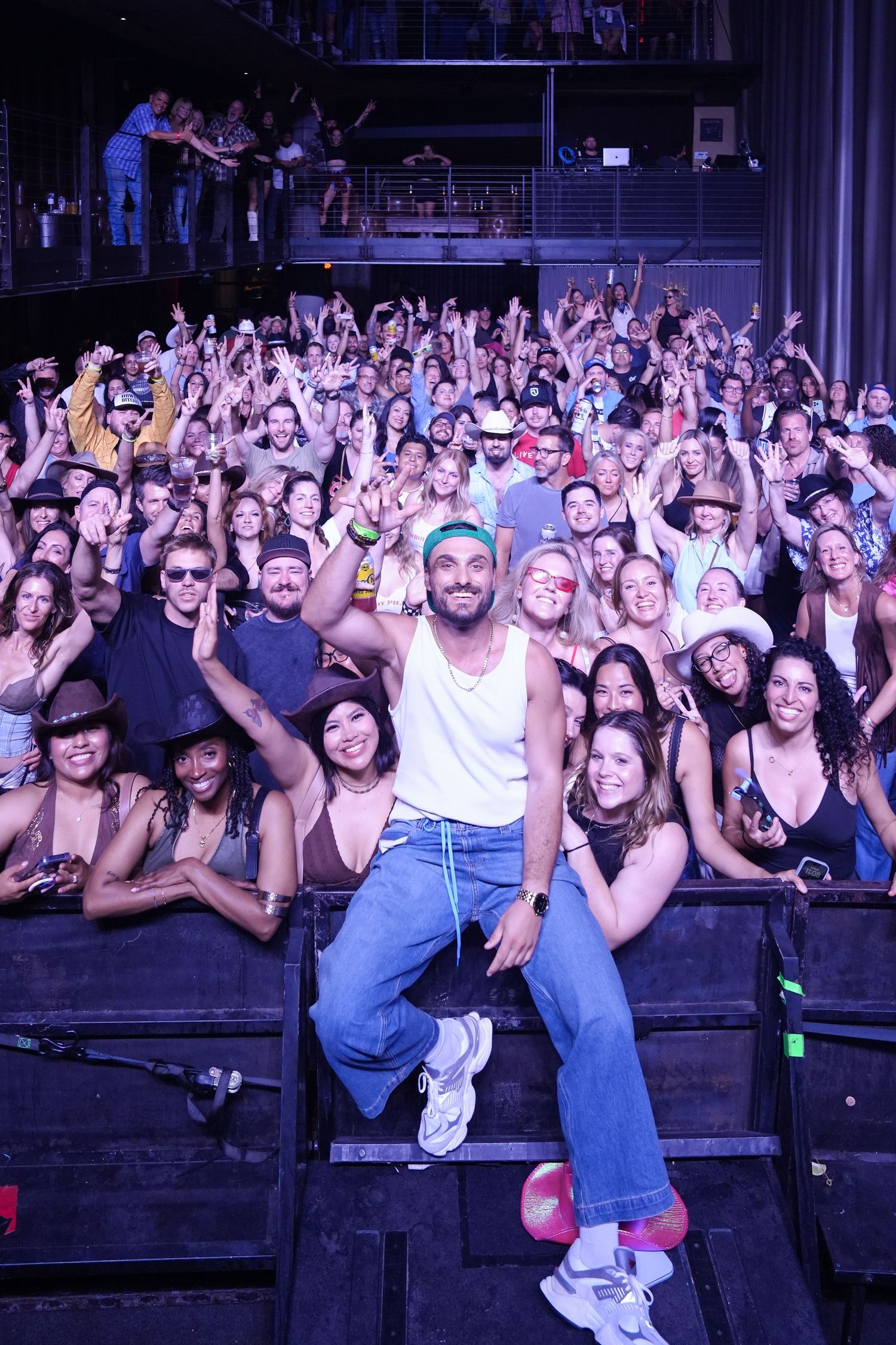 Excited concert crowd cheering and raising their hands at a nighttime music venue with purple lighting.