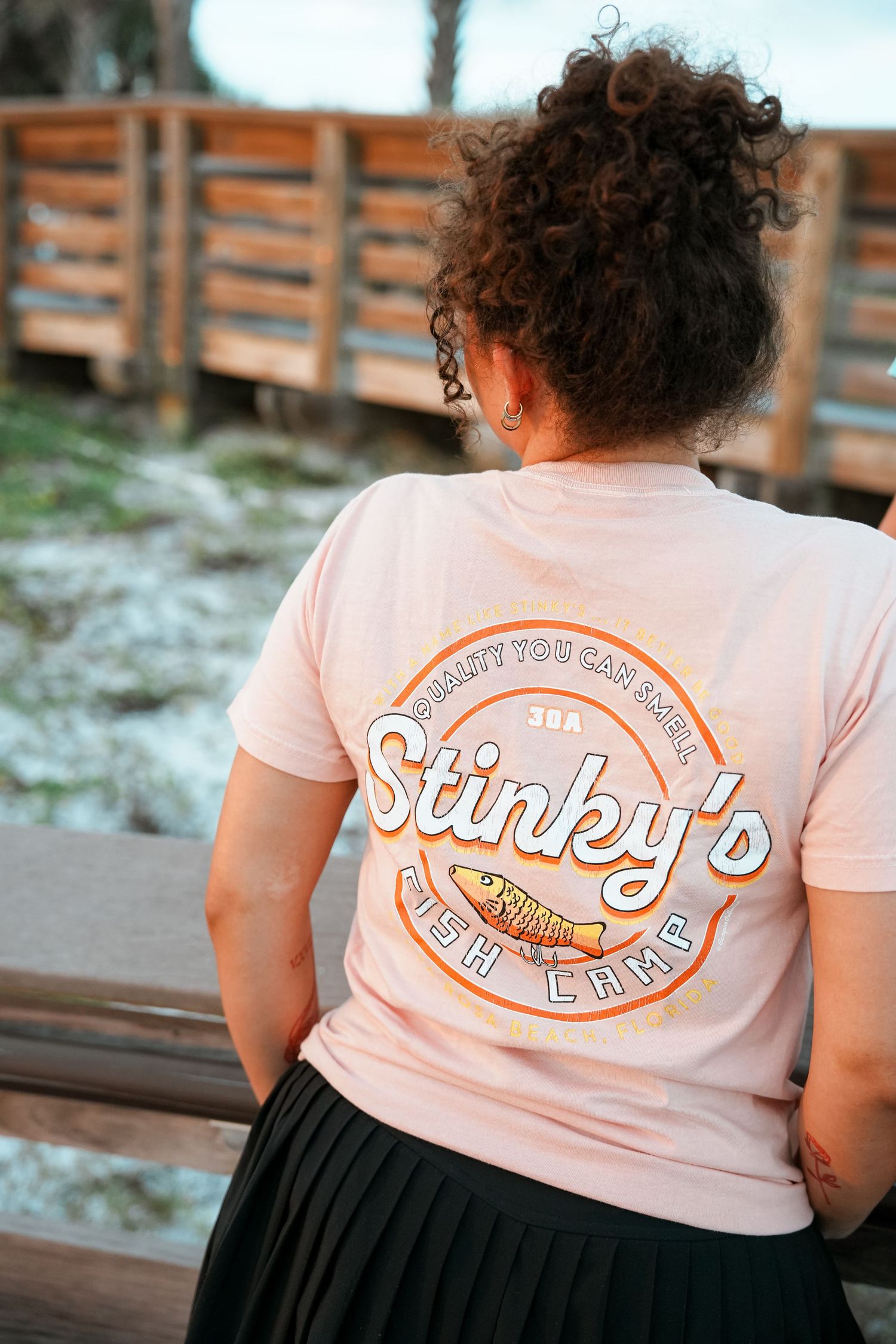 A person wearing a peach-colored t-shirt with 'Stinky's Fish Camp' logo sitting on a wooden bench.