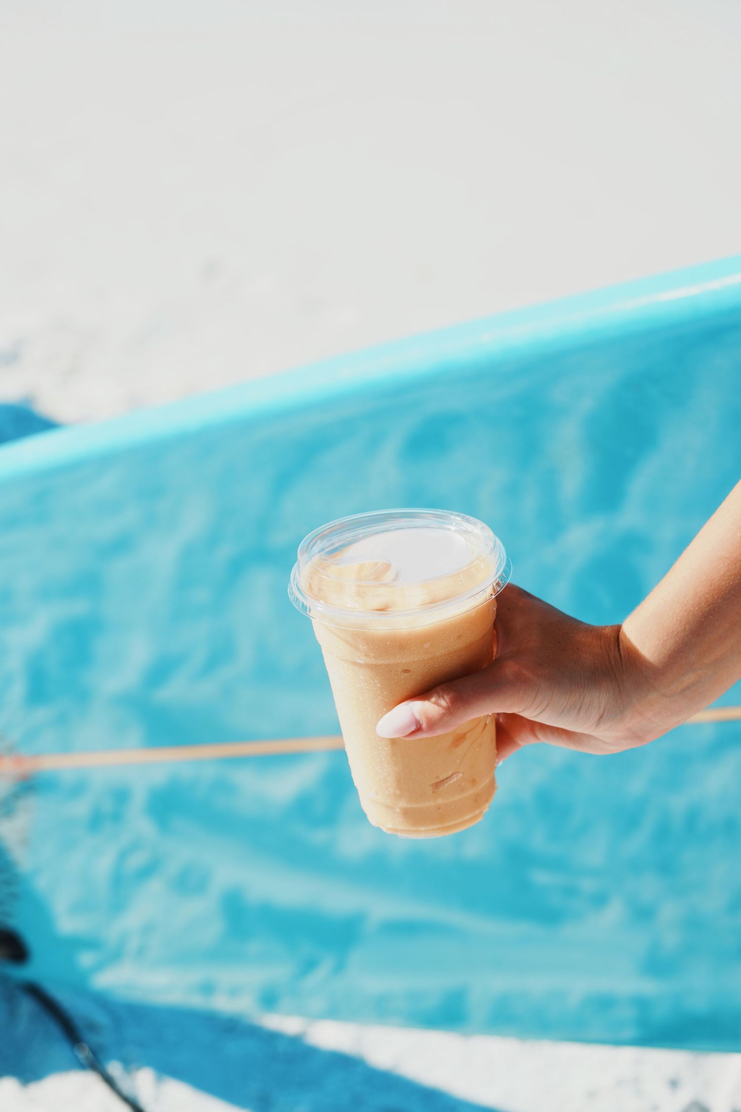 A hand holds a clear plastic cup of creamy orange smoothie against a bright turquoise blue background.