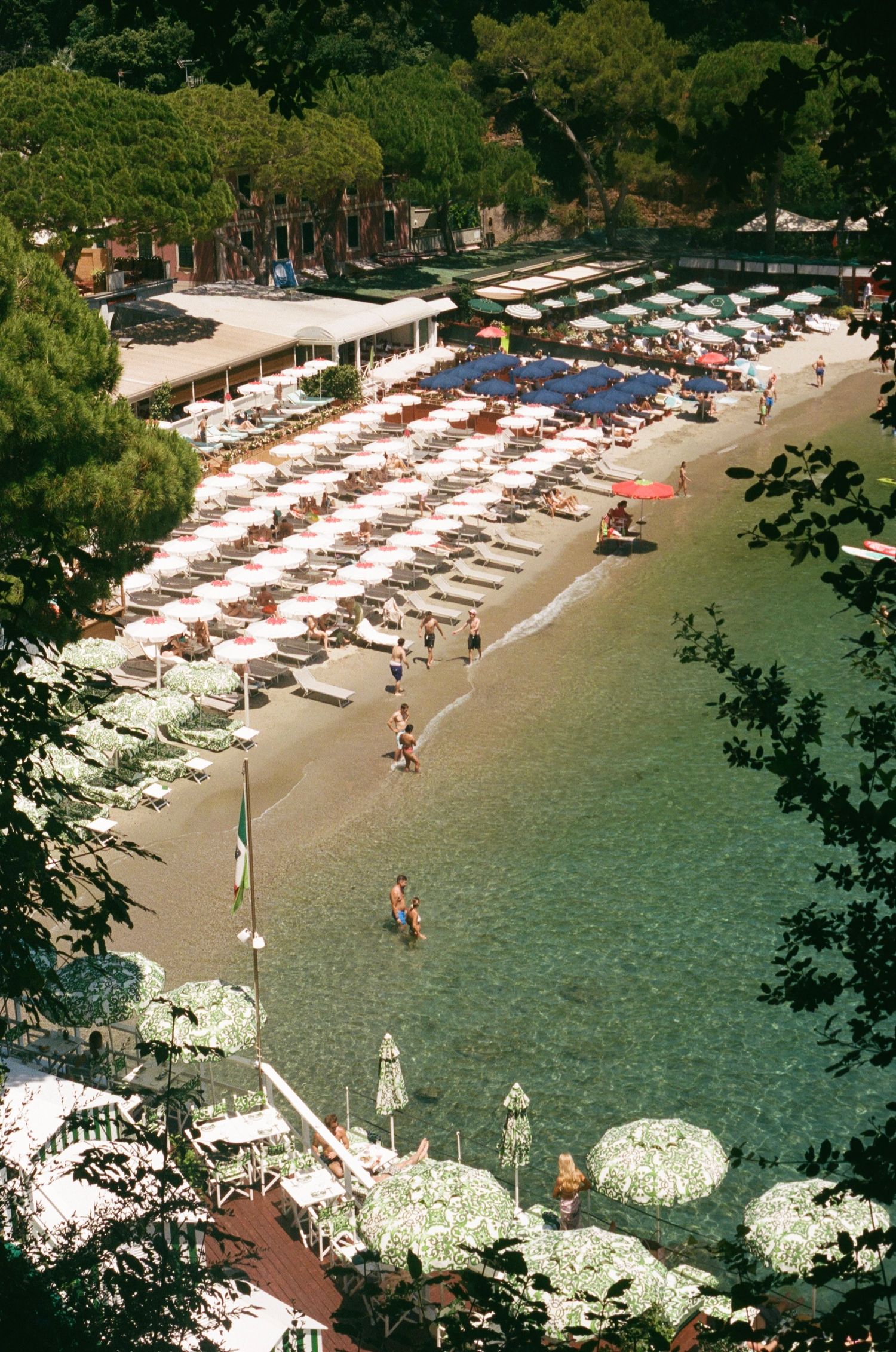 Aerial view of a scenic beach resort with colorful umbrellas, lounge chairs, and clear turquoise water surrounded by lush trees.