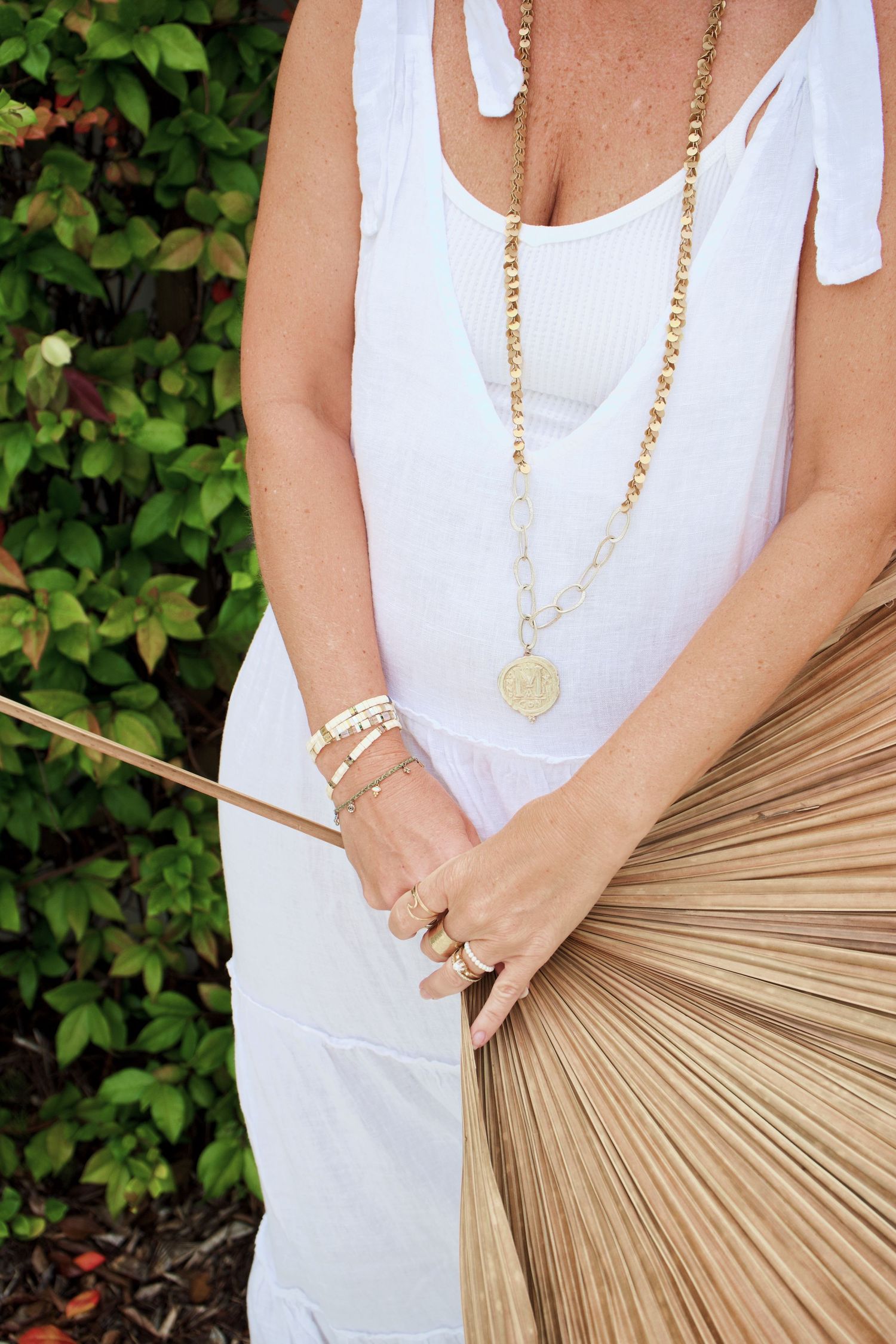 Woman in white sundress with gold necklace and bracelets holds a tan woven fan against green foliage background.