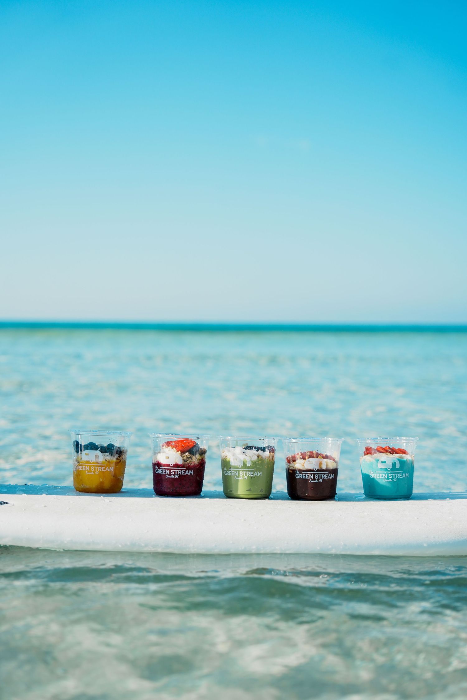 Colorful smoothie bowls topped with berries and nuts lined up on a white ledge against turquoise tropical ocean waters.