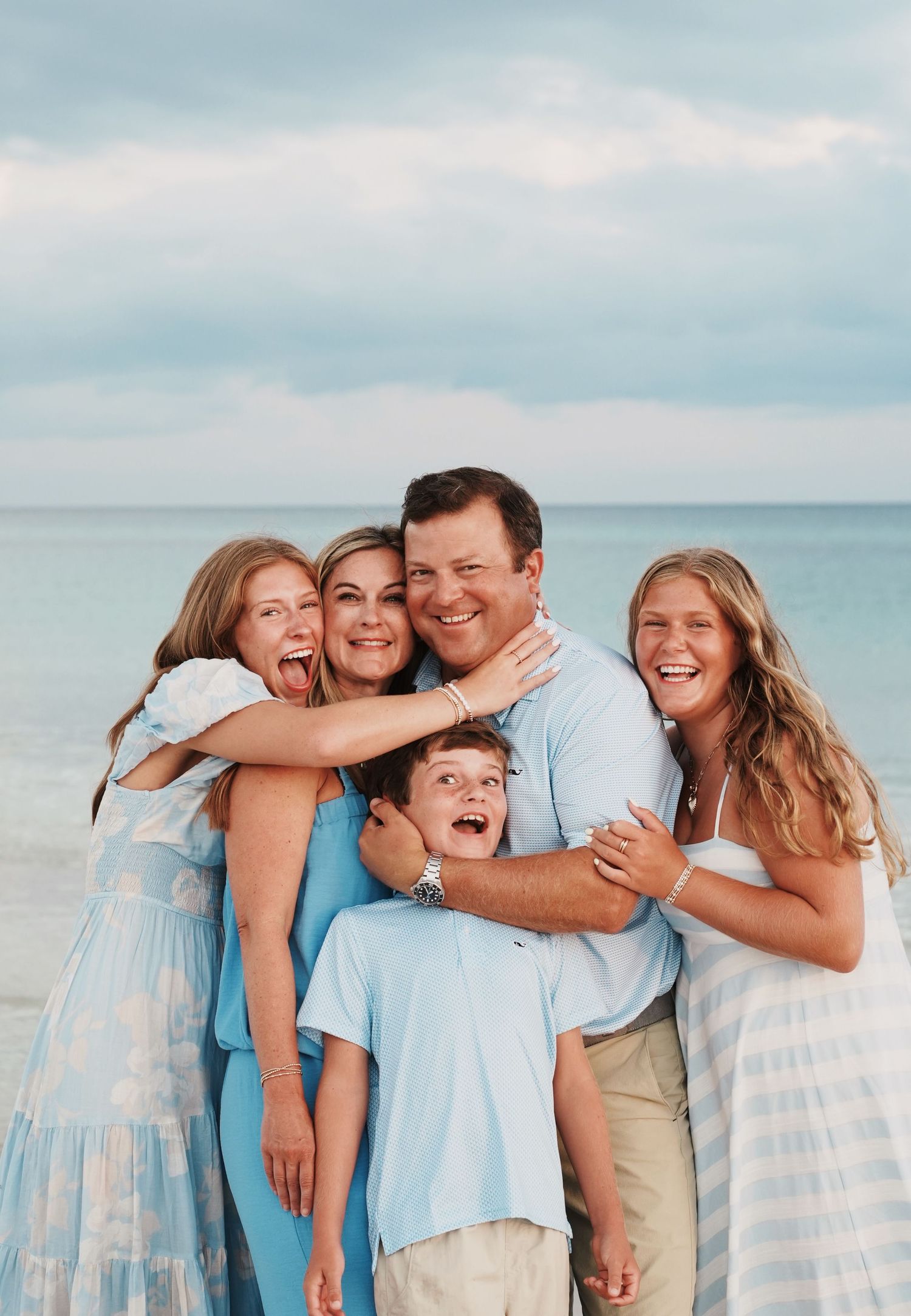 A playful casual beach photo session with people in light blue and white outfits smiling and embracing on a cloudy day.