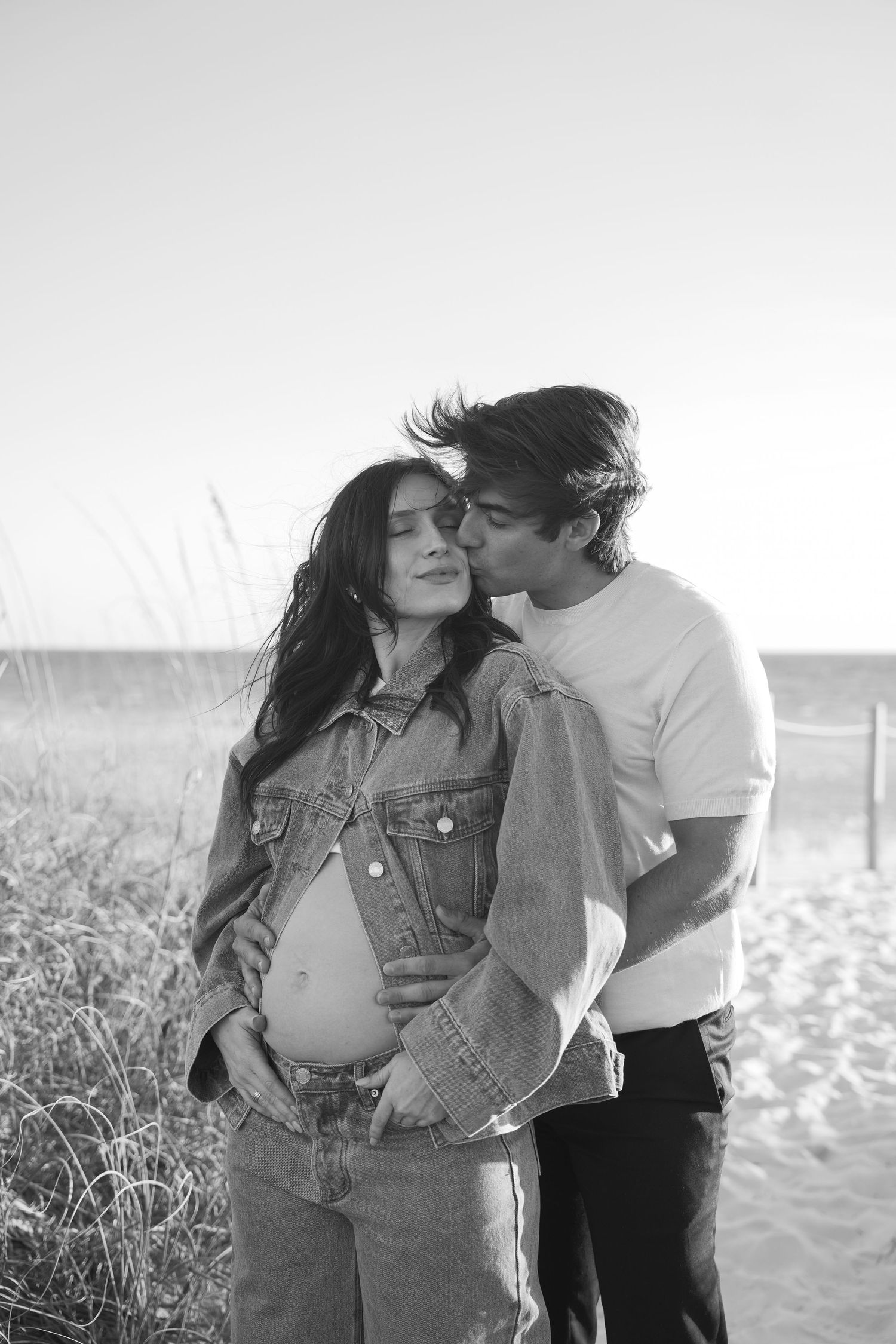 Black and white romantic beach photoshoot series showing a couple in denim embracing on a sandy shoreline.
