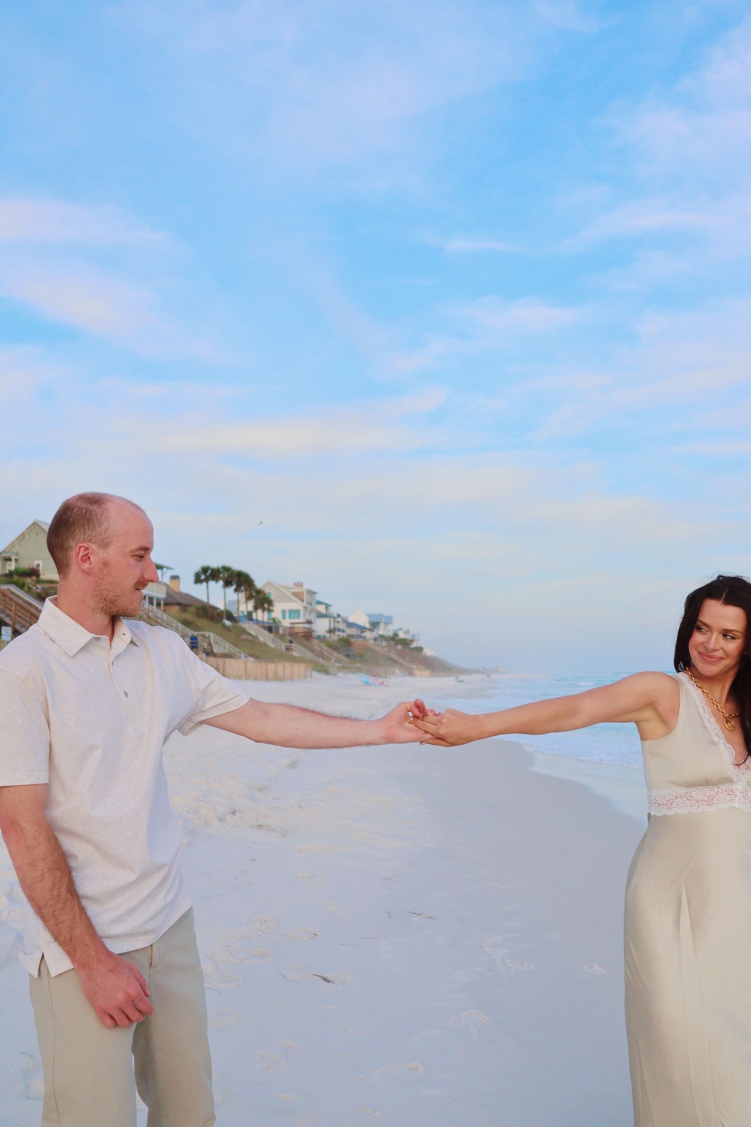 Couple in matching white and cream outfits holding hands on a sandy beach.