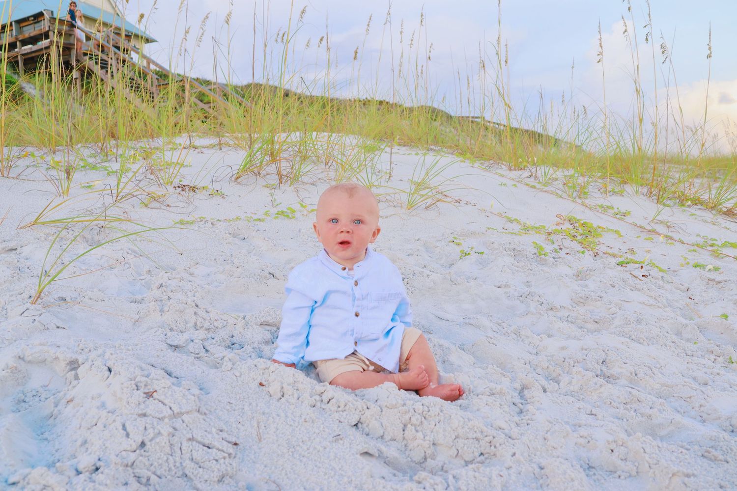 Baby in blue outfit sits on white sand beach.