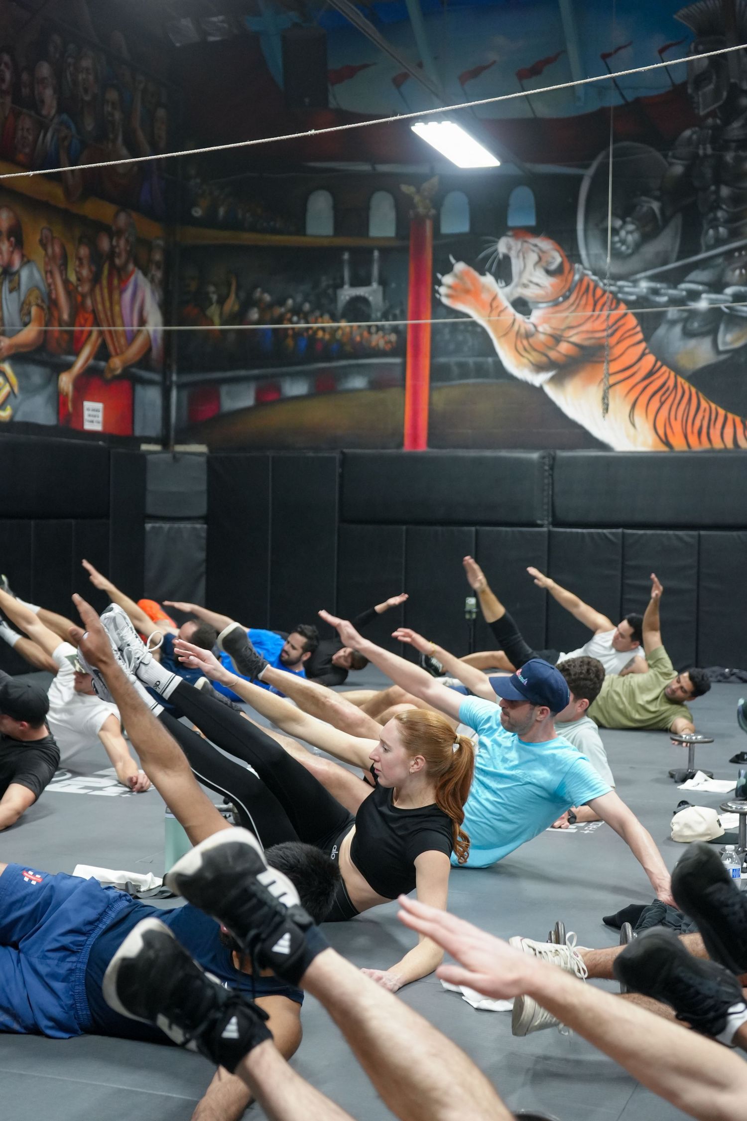 Group fitness class participants lying on mats performing synchronized stretching exercises in a gym with colorful wall murals.