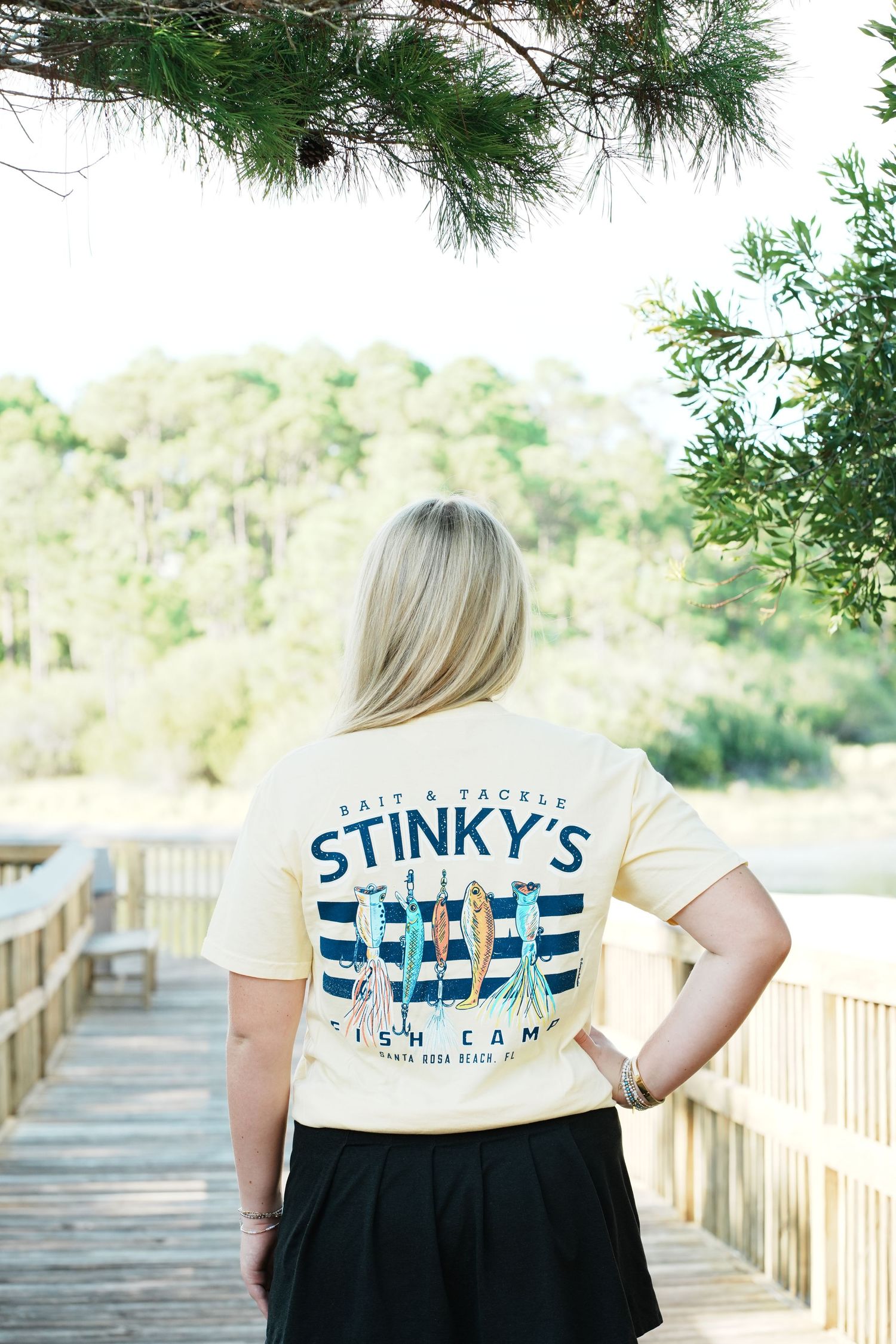 Person wearing a beige Stinky's Fish Camp t-shirt stands on a wooden boardwalk surrounded by pine trees.