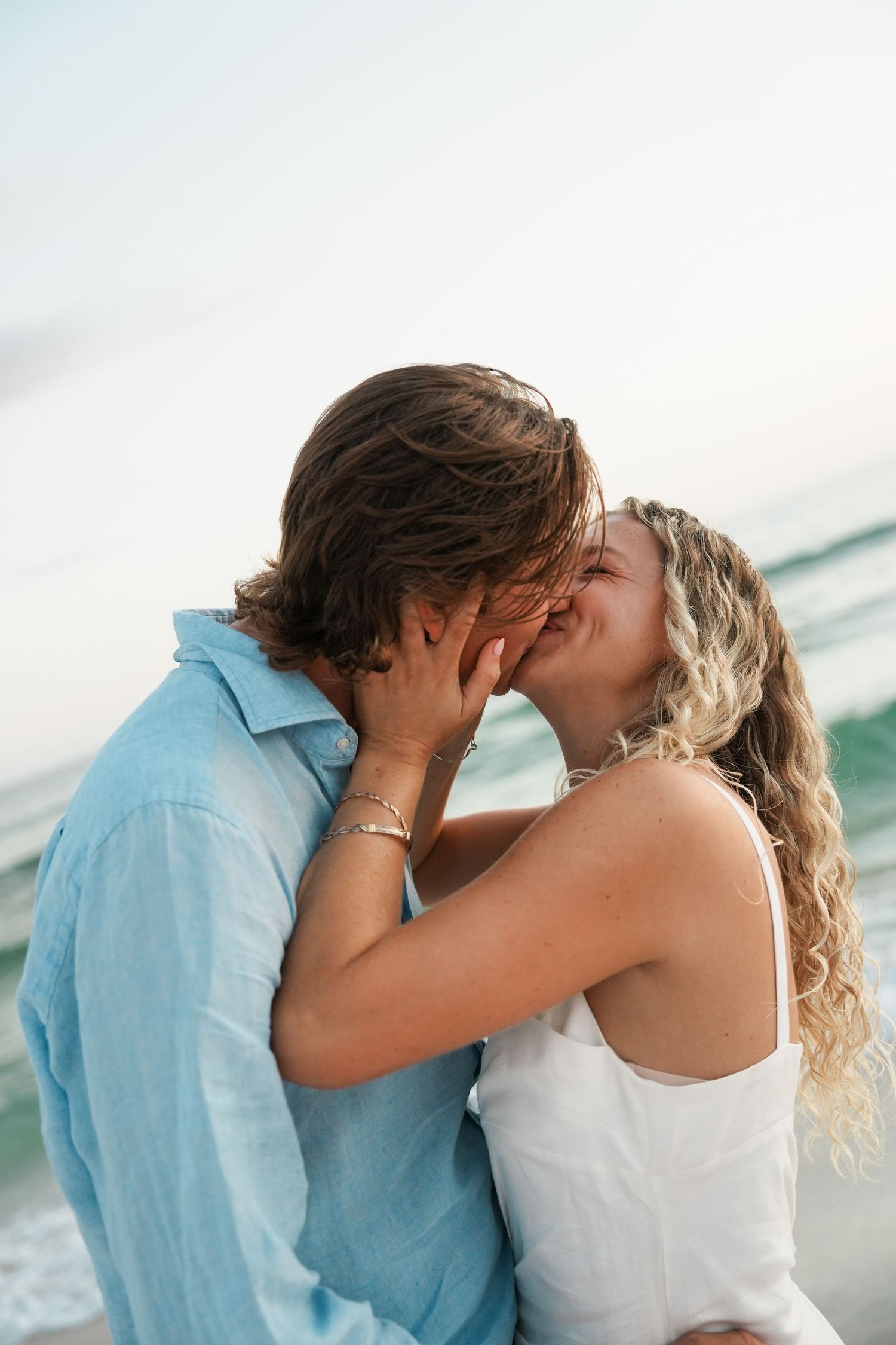 A romantic couple embraces and kisses on a beach during sunset with ocean waves in the background.