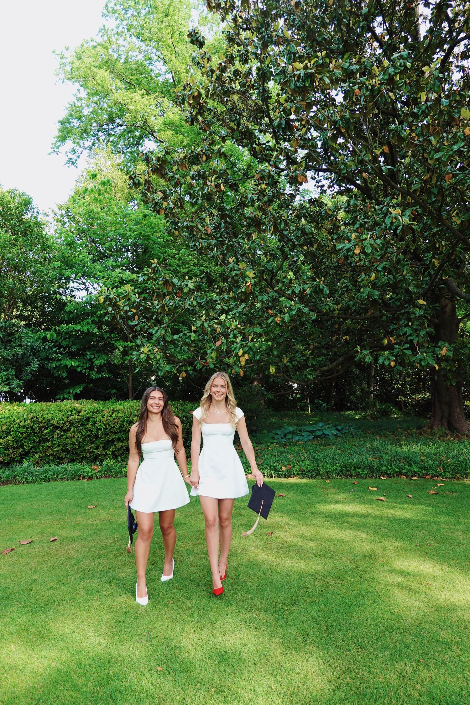 Two people in white summer dresses walking together on a manicured lawn with lush green trees in the background.