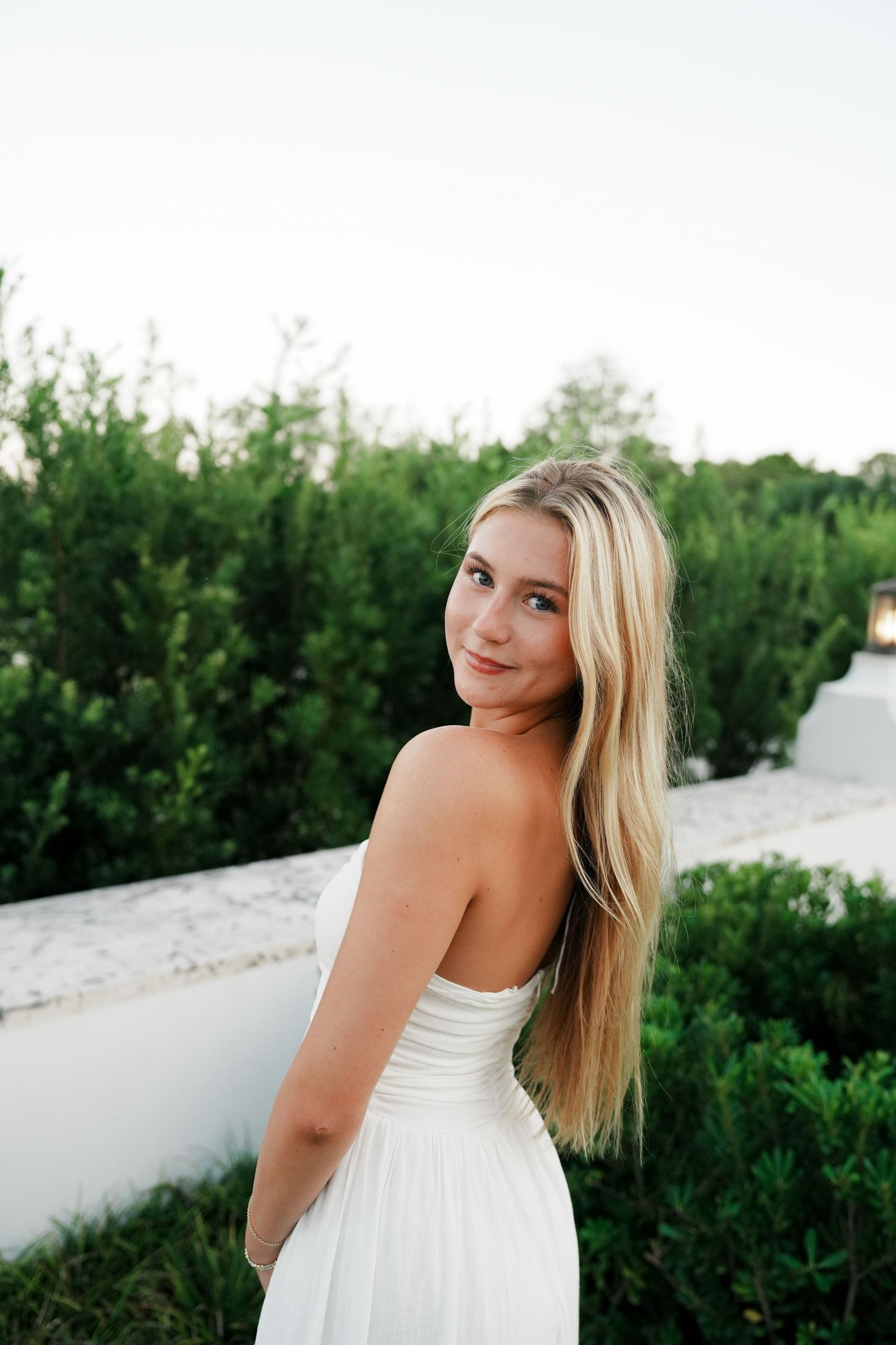 A person in a white strapless dress poses outdoors against a backdrop of green bushes and a white wall.