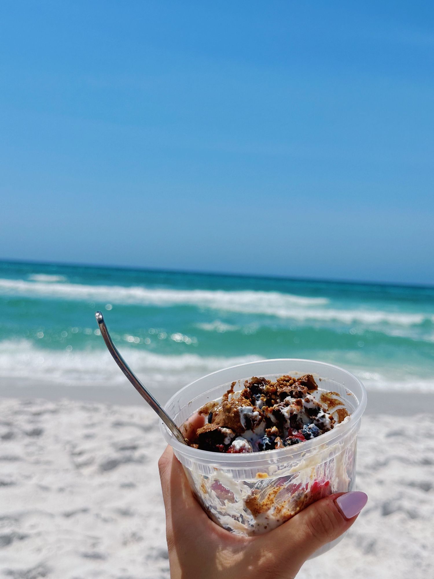 Ice cream dessert with toppings held against a scenic beach background with turquoise waves and white sand.