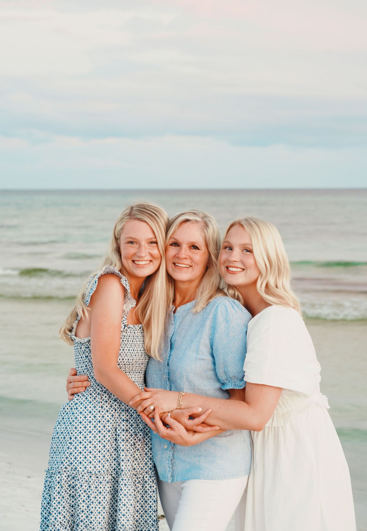 Three people in light colored clothing embrace and smile together on a beach with the ocean visible in the background.