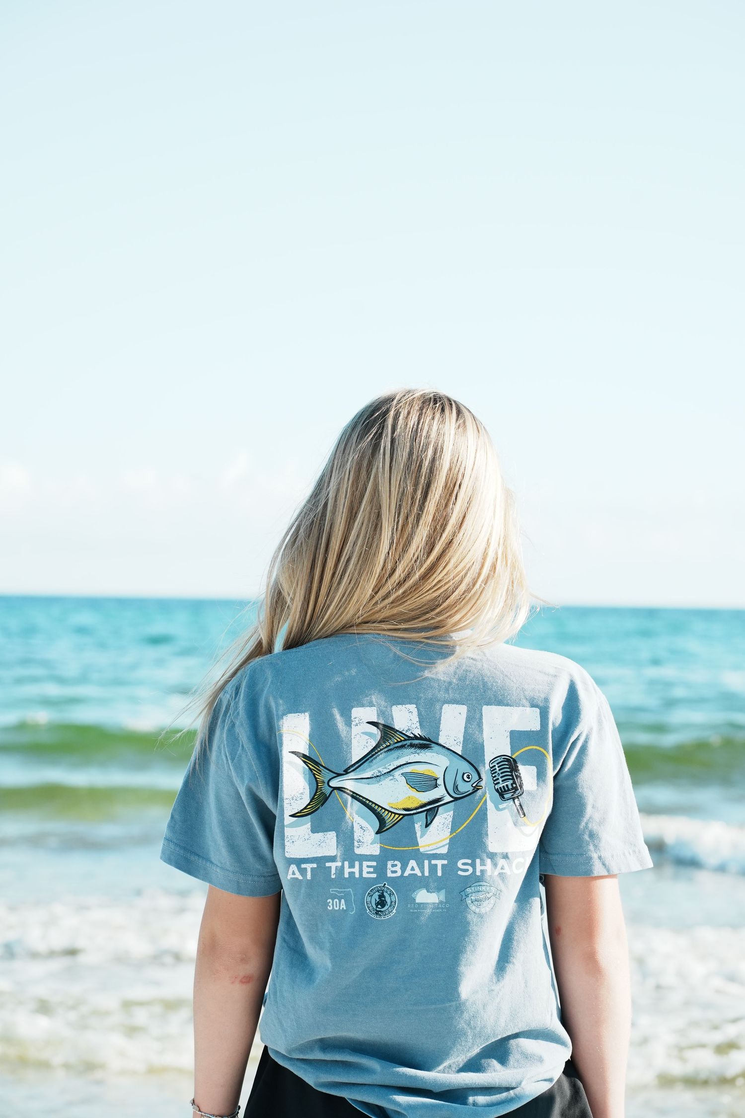 Person wearing light blue t-shirt with fishing logo design standing on beach with ocean waves in background.