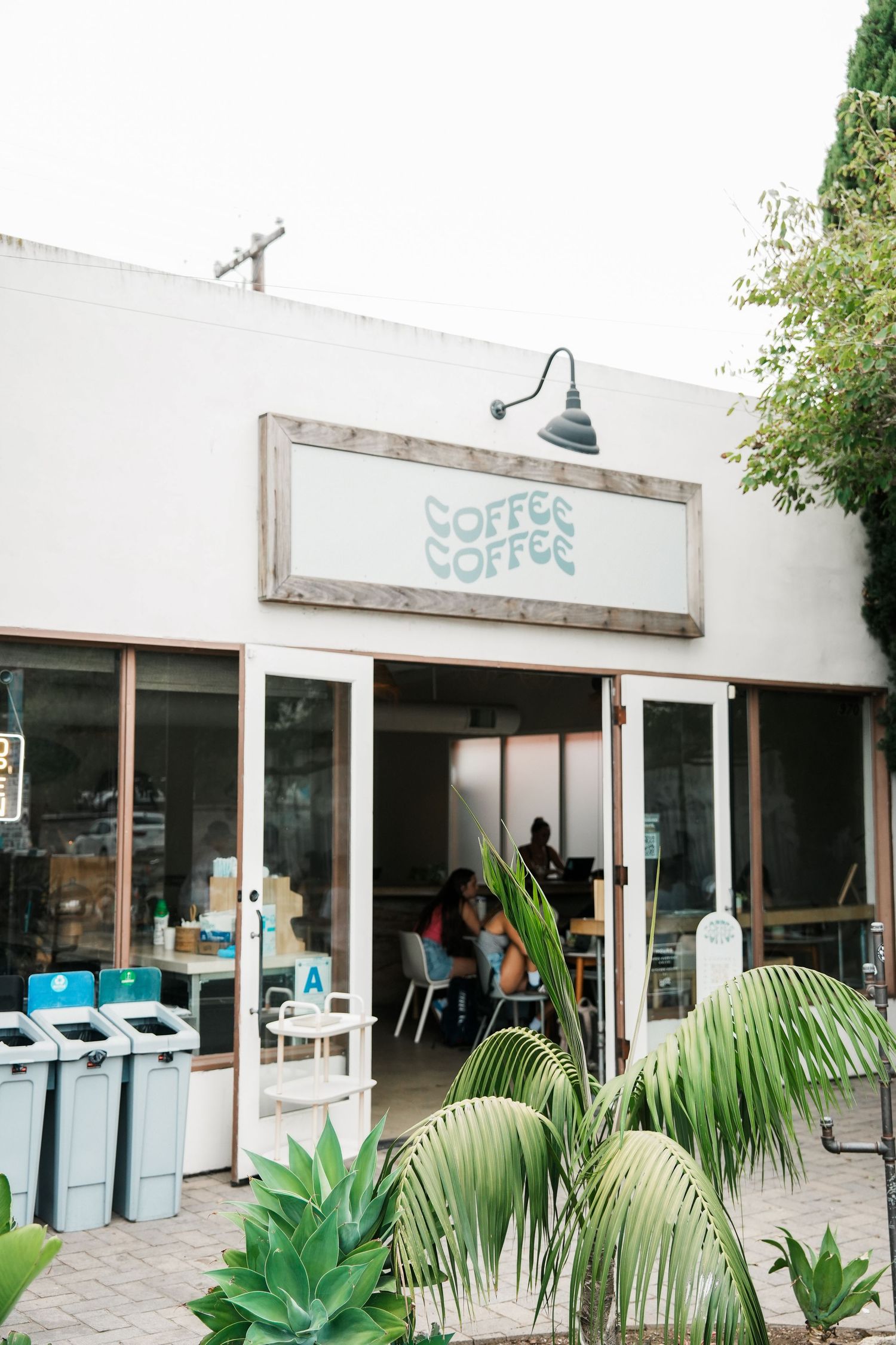 Modern white coffee shop exterior with tropical plants and outdoor seating area.