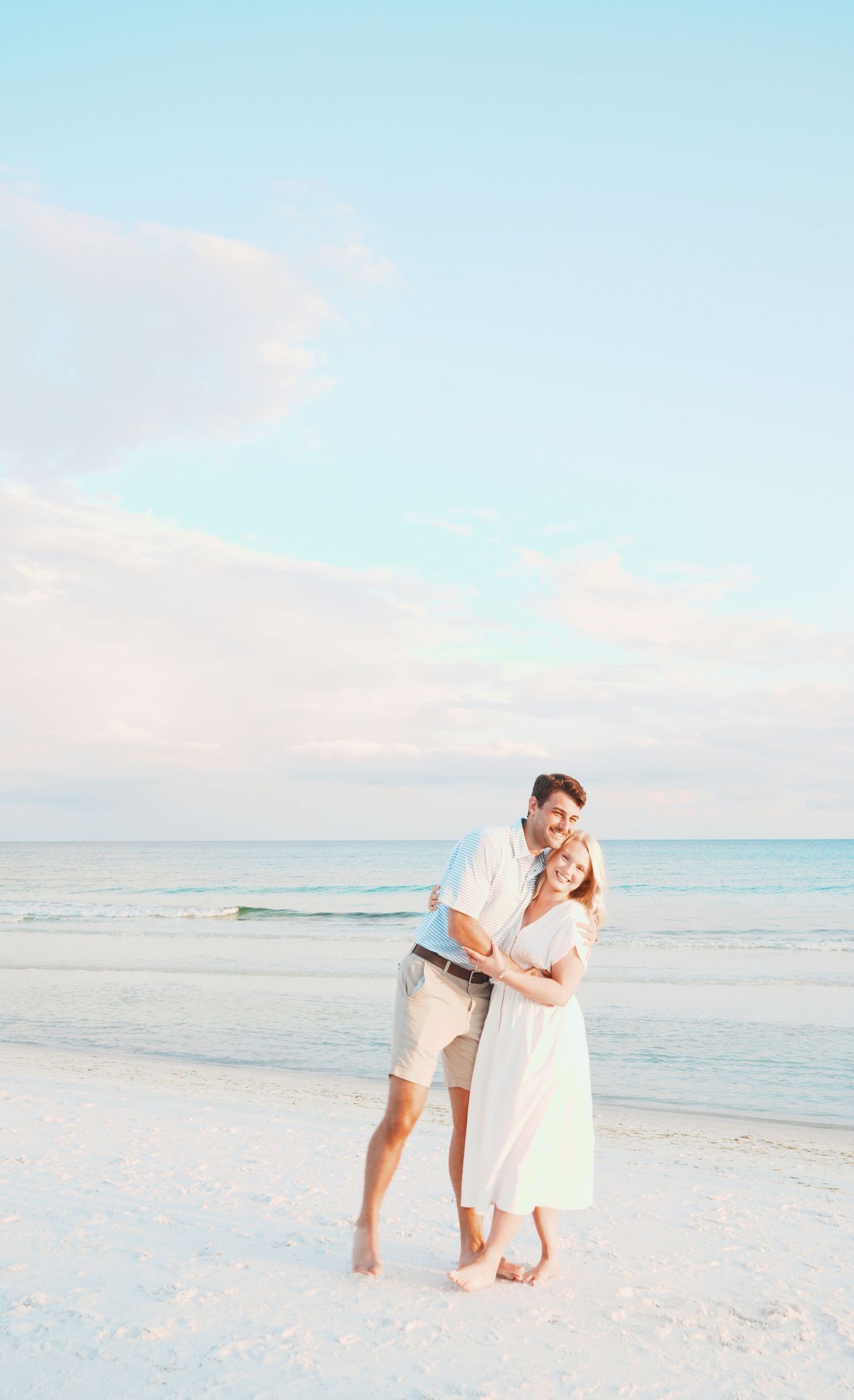 A romantic couple in white outfits share tender moments together while walking along a dreamy pastel beach at sunset.
